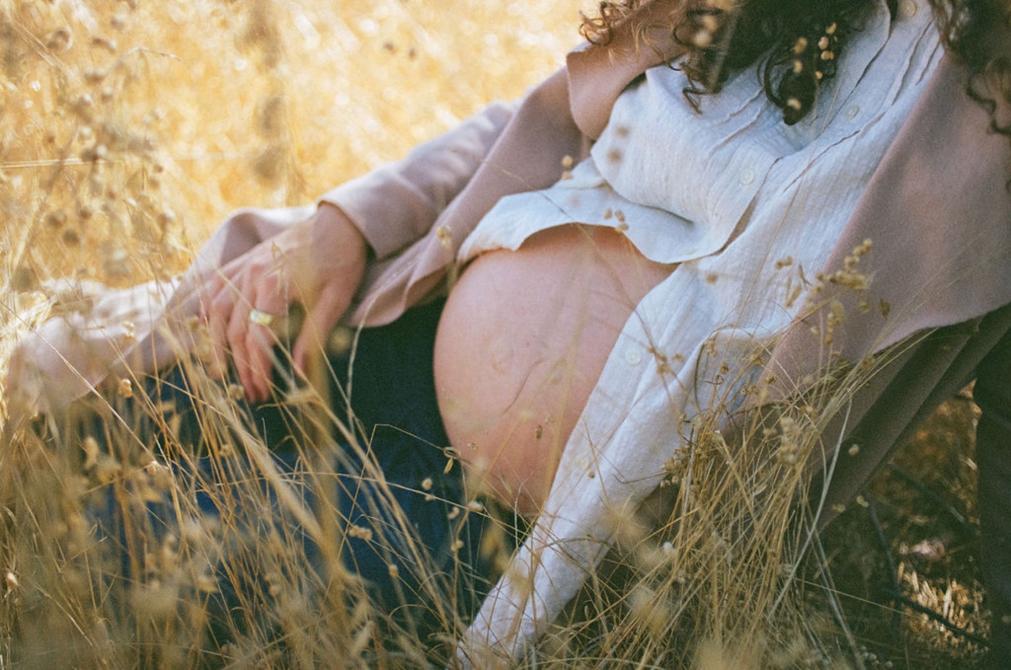 Pregnant woman in white linen sits cradled by golden grasses, her bare belly glowing in warm afternoon light while her partner's arm tenderly wraps around her shoulders