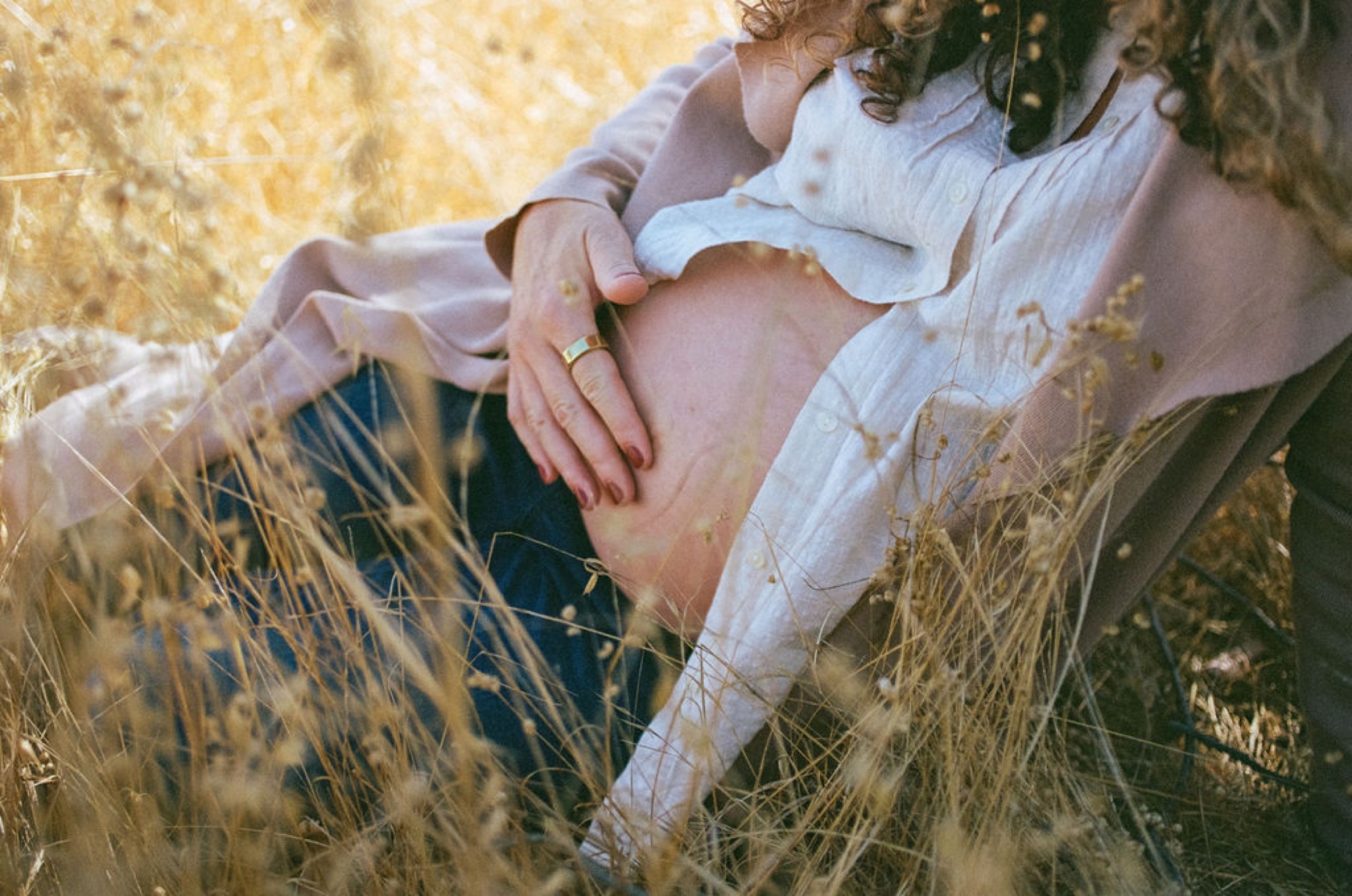 Pregnant woman in white linen shirt cradles her belly while seated in golden dried grasses, hands gently framing her abdomen with tender reverence