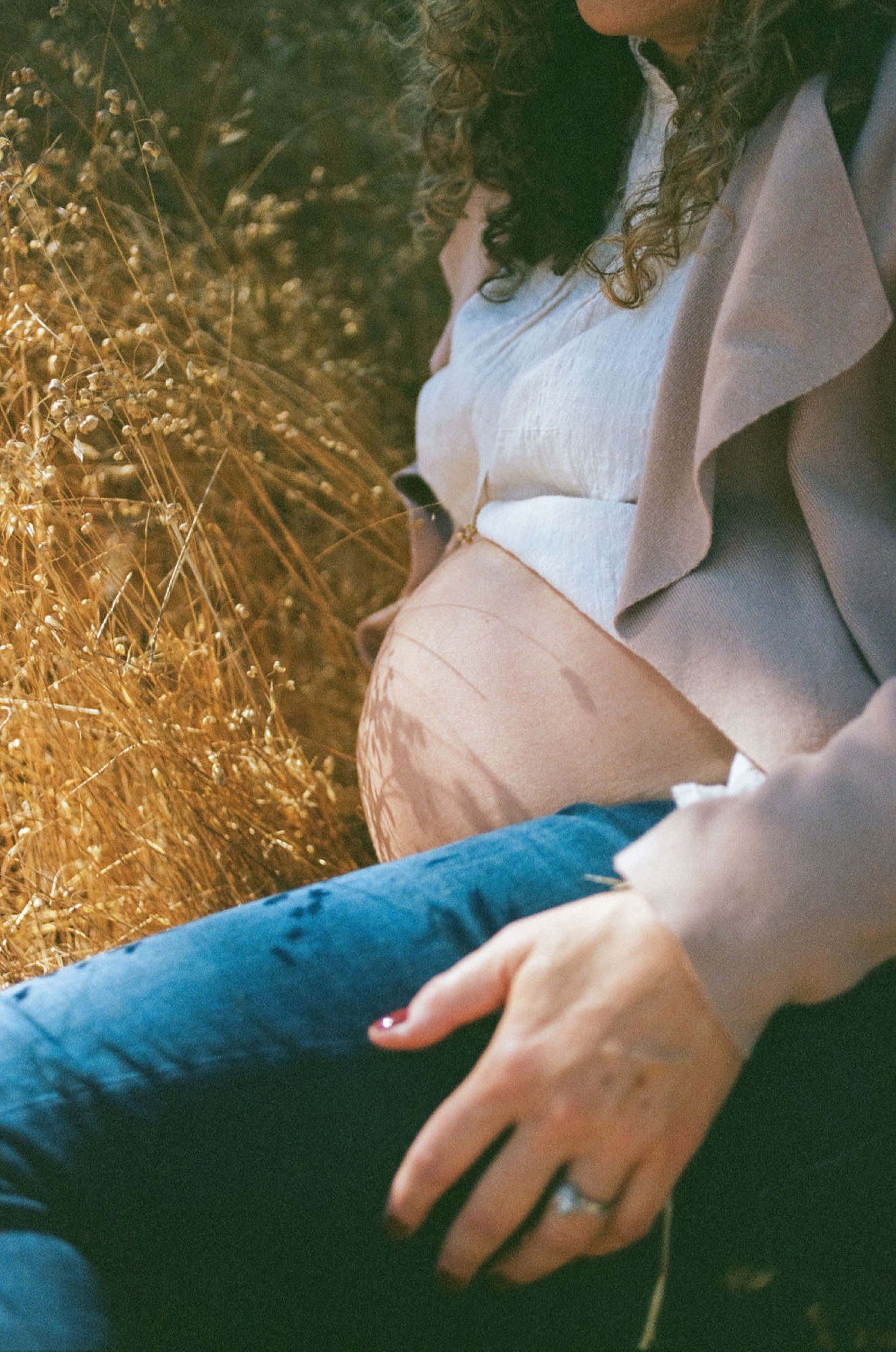 Pregnant woman in white crop top cradles her belly while sitting among golden dried grasses, her partner's hand resting on her leg in tender connection