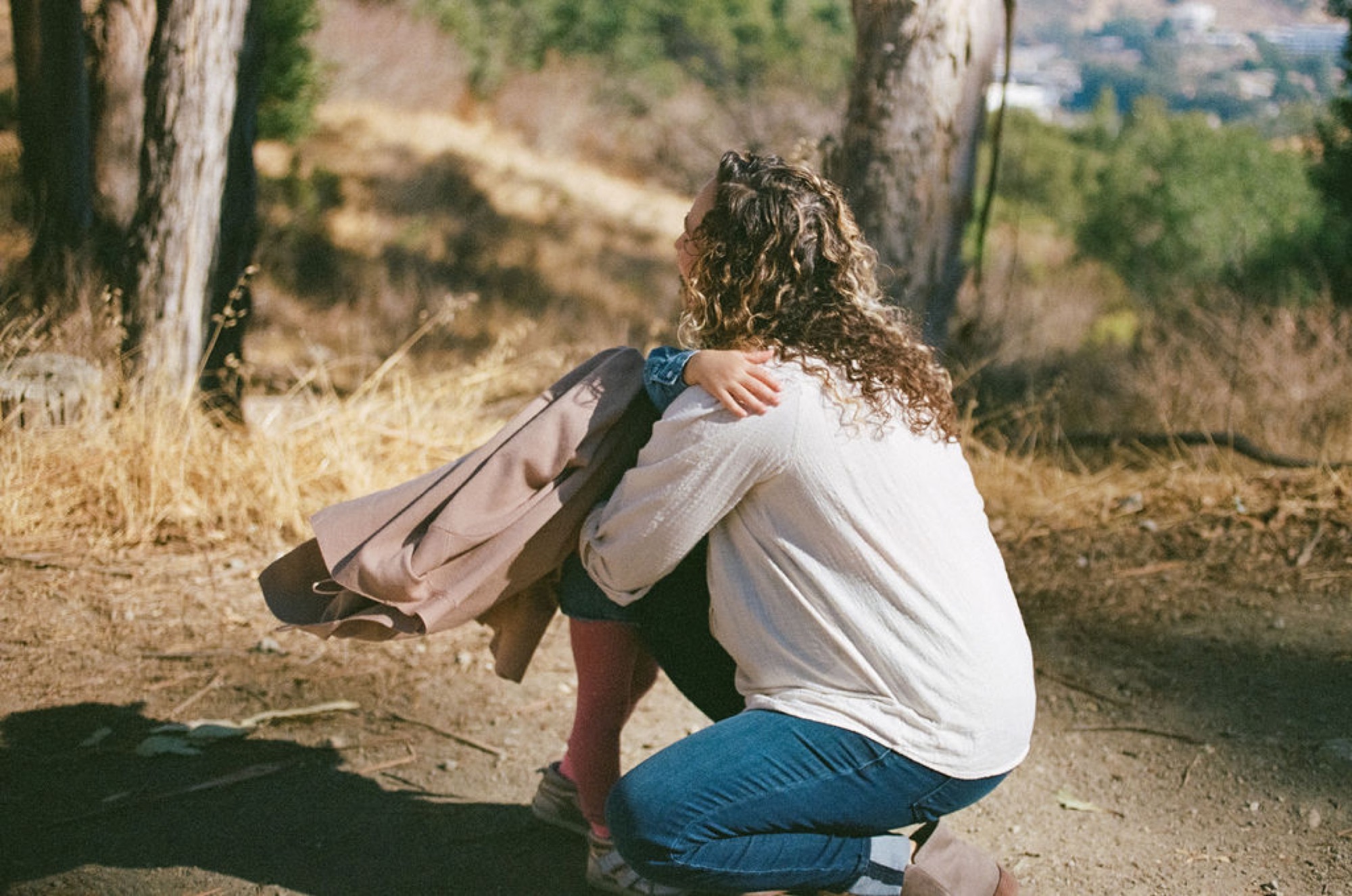 Pregnant woman crouches on a dusty trail between eucalyptus trees, cradling her belly with a contemplative gaze toward the distant hillside