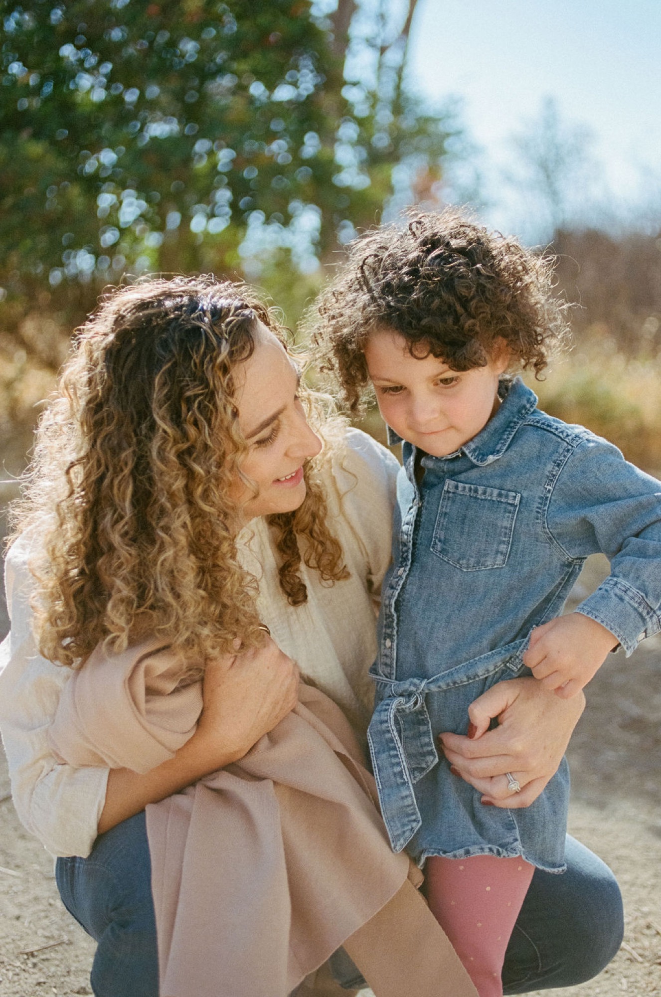 Woman in neutral tones cradles her young child in denim on a sunlit path, both gazing downward with gentle expressions
