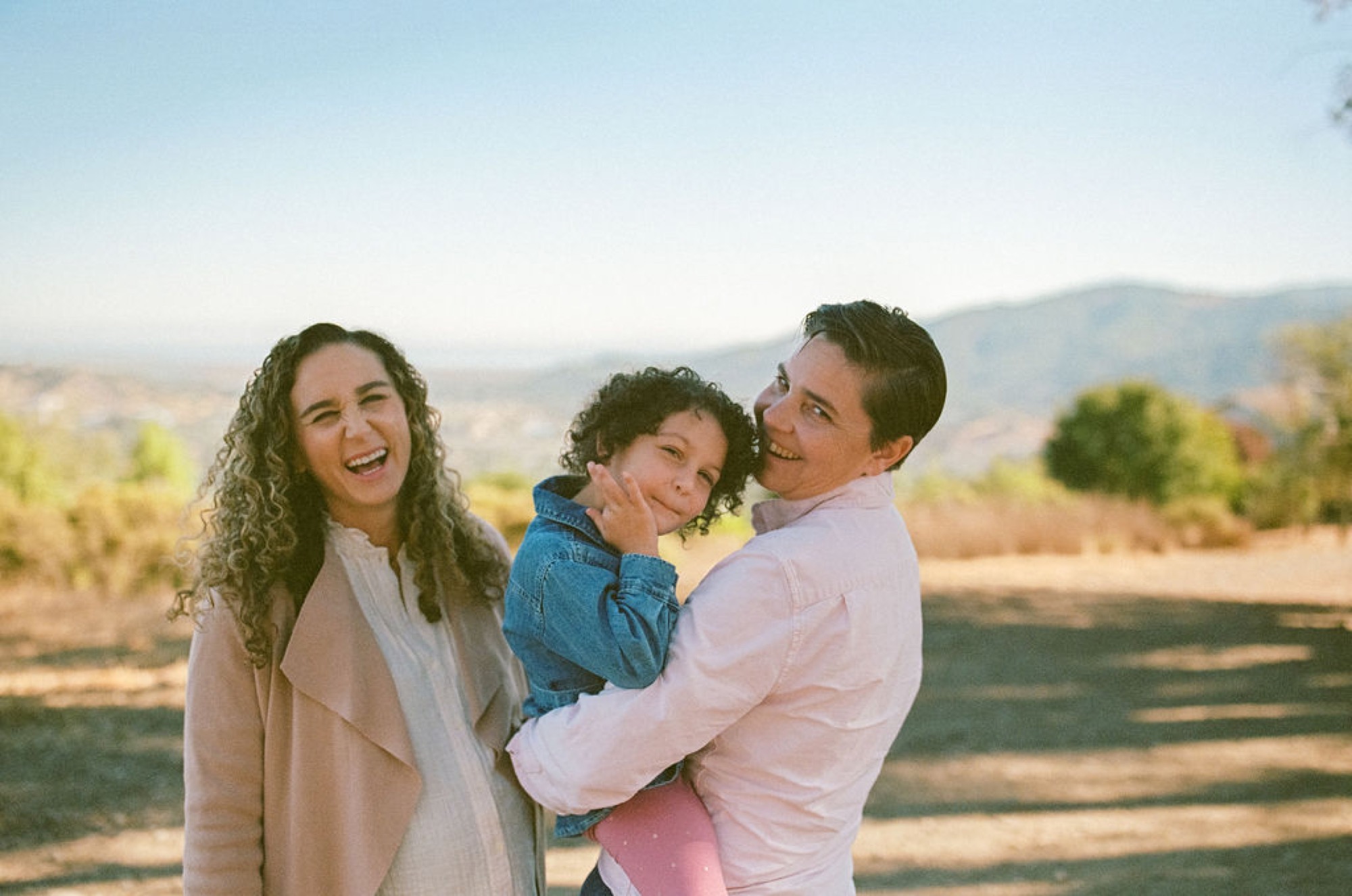 A family of three laughs together on a sun-drenched hillside, one mom holding their young child while the other beams beside them, golden afternoon light washing across the rolling landscape
