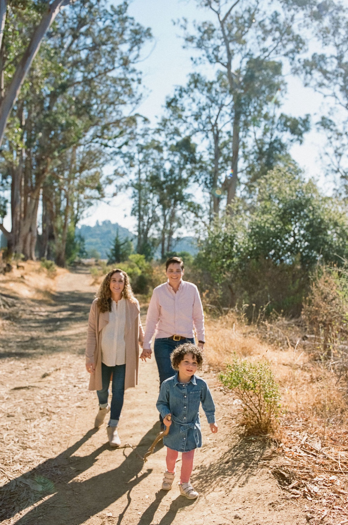 A young family walks together down a sun-dappled dirt trail, with a toddler leading ahead while parents hold hands behind, mountains visible through the tall eucalyptus trees