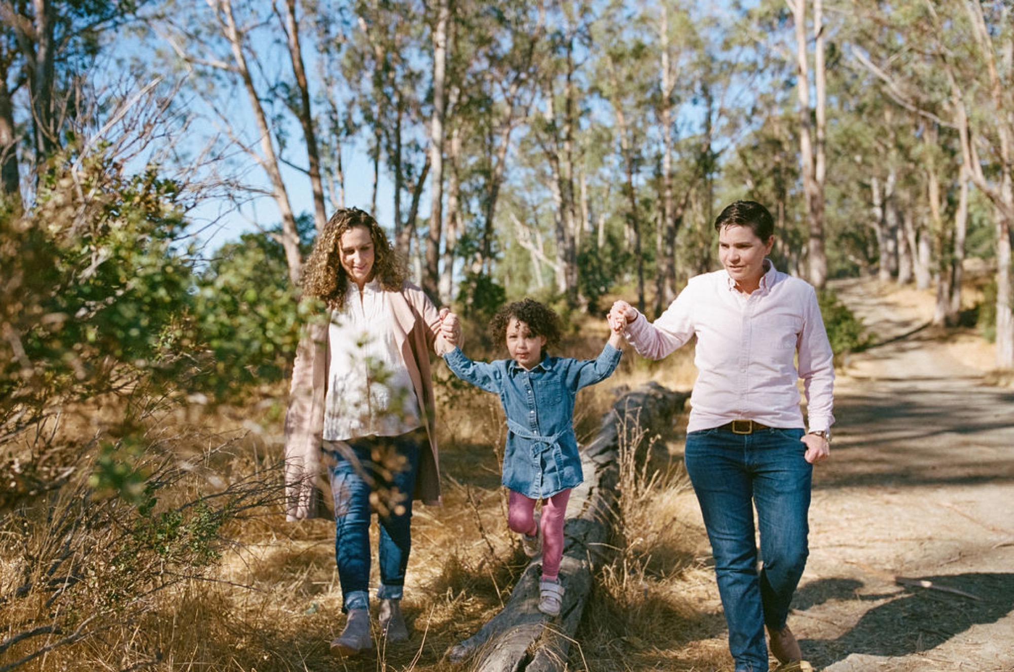 A family of three walks hand-in-hand down a sunlit dirt trail through tall eucalyptus trees, with the child lifted mid-stride between the two adults