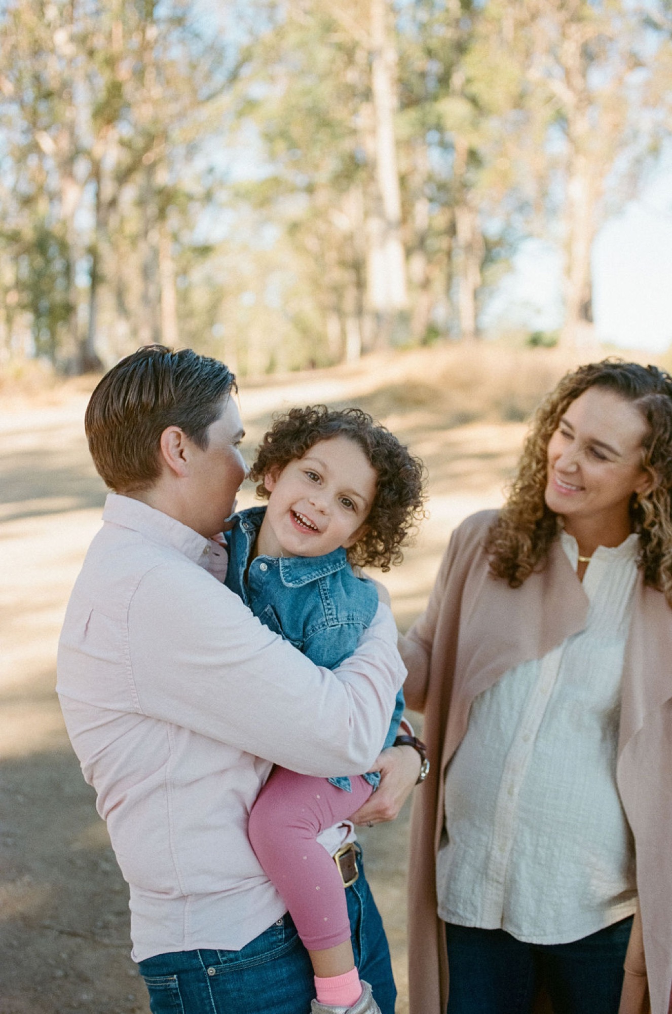 A man holds his laughing curly-haired daughter while his wife smiles beside them on a sun-dappled tree-lined path
