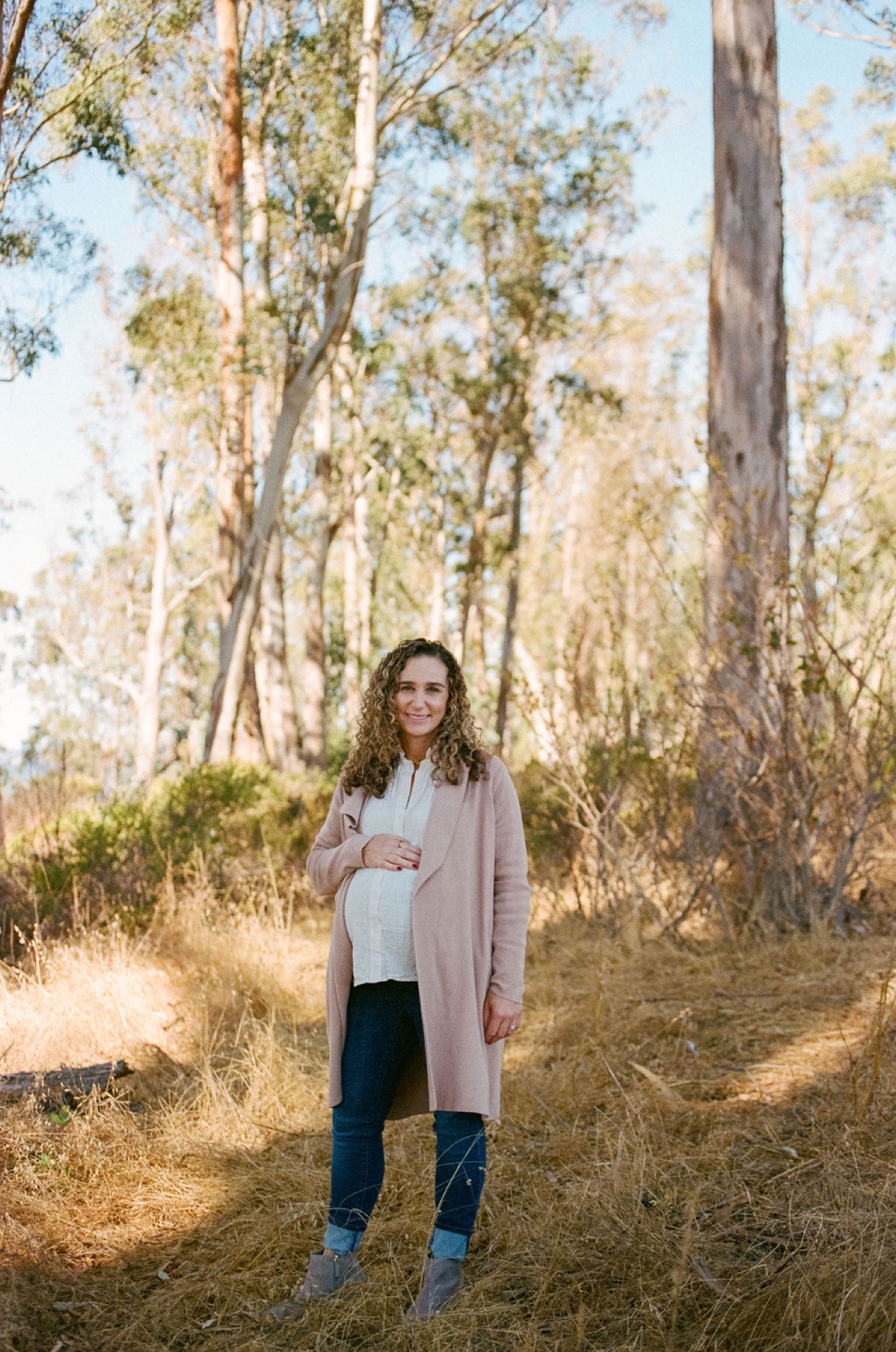 Pregnant woman with curly hair smiles warmly while cradling her belly, standing on a sunlit forest trail lined with towering eucalyptus trees