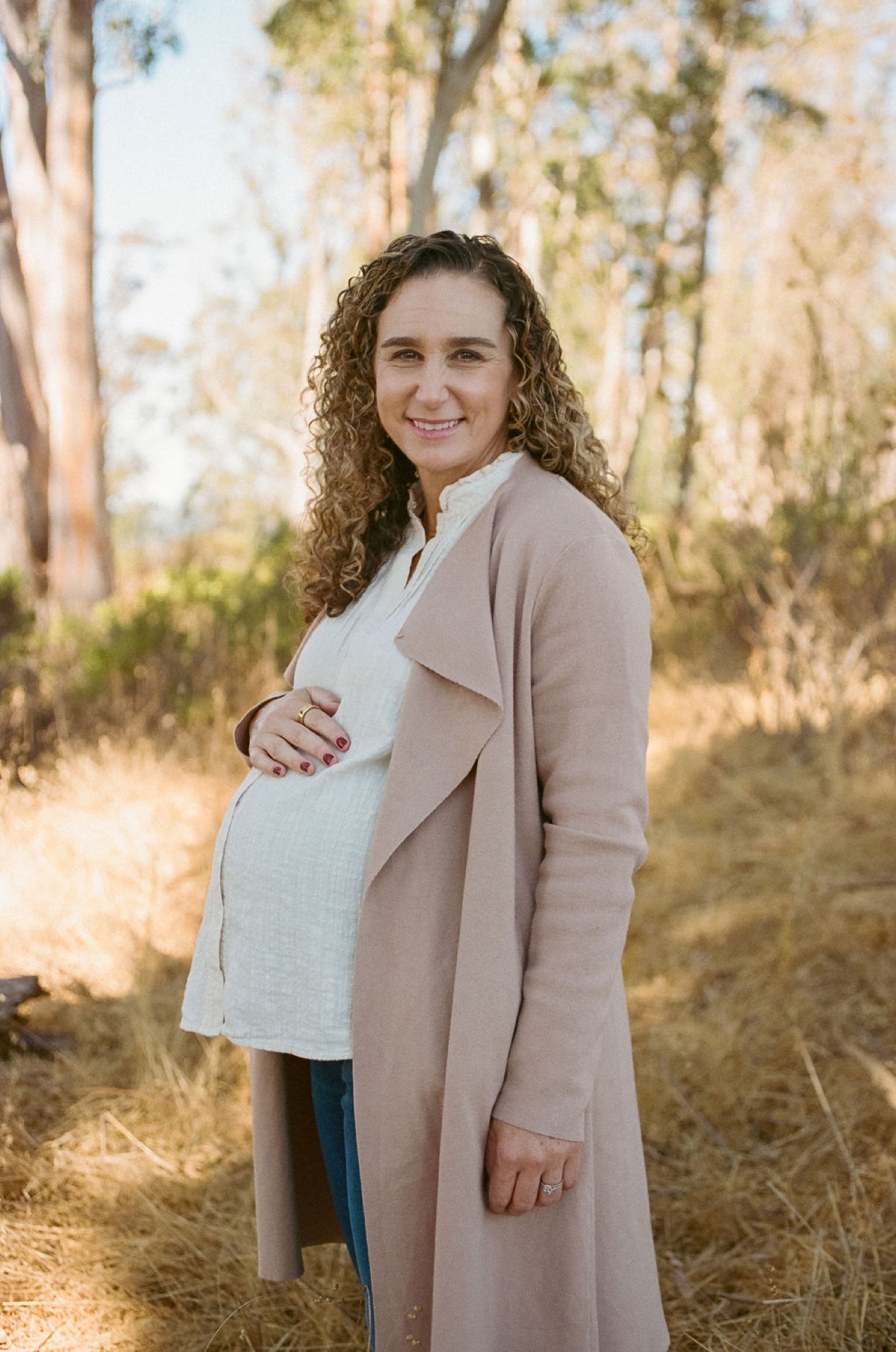 Expectant mother smiles warmly at camera while cradling her belly, wearing a cream cardigan over a white top amid tall eucalyptus trees