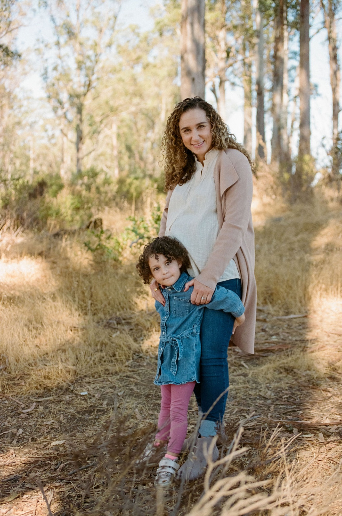 Pregnant woman and young daughter walk hand-in-hand down a sunlit forest trail, both smiling warmly at the camera