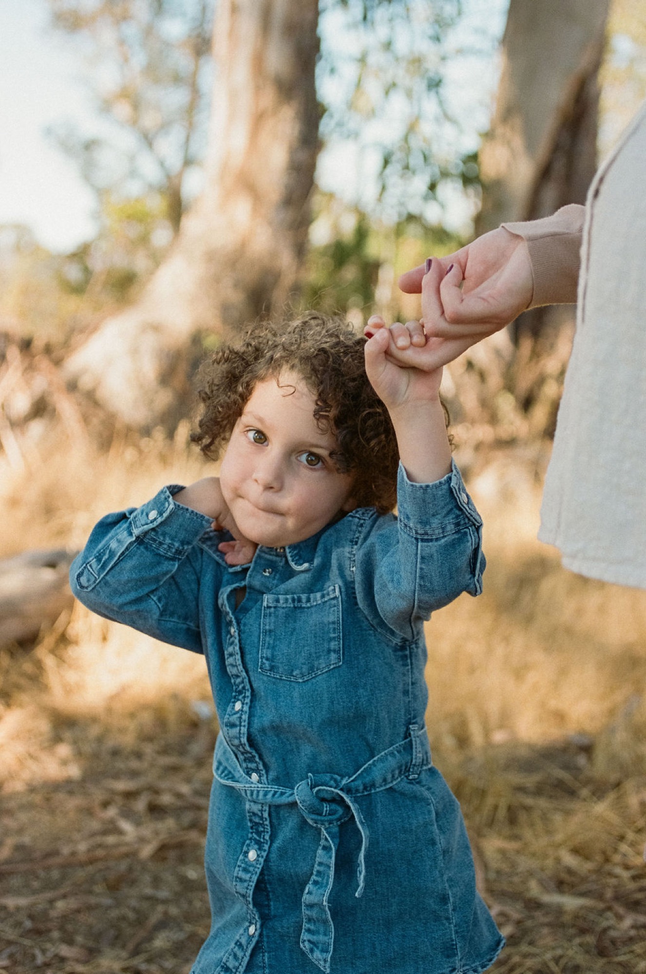 Toddler in denim dress gazes upward while holding an adult's hand beneath blooming white flowers and golden hillside grasses