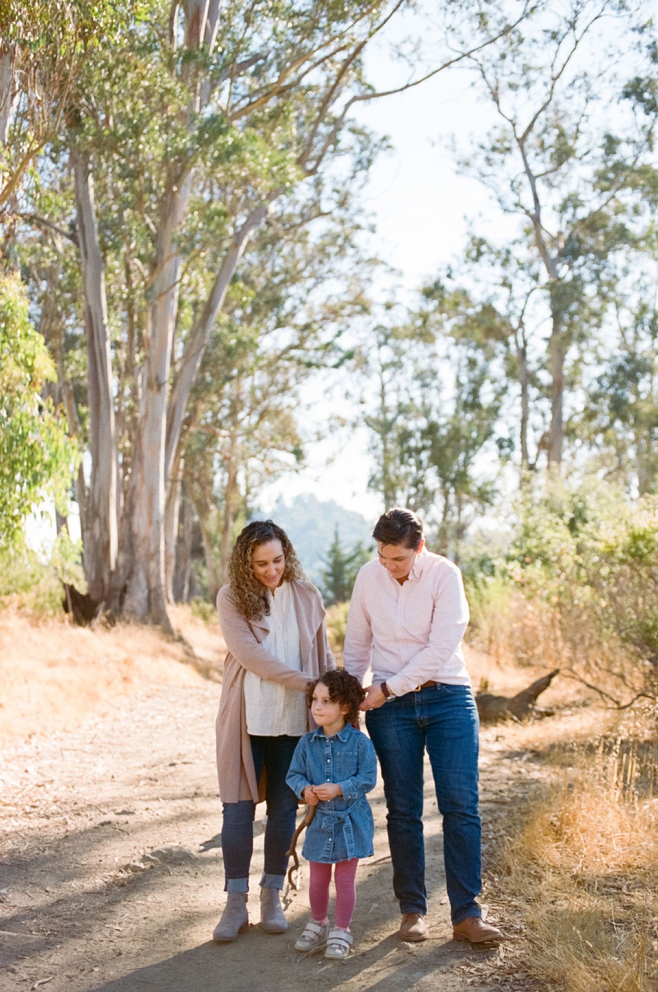Two women and their young child walk hand-in-hand down a sunlit dirt path lined with towering eucalyptus trees, mountains visible in the distance