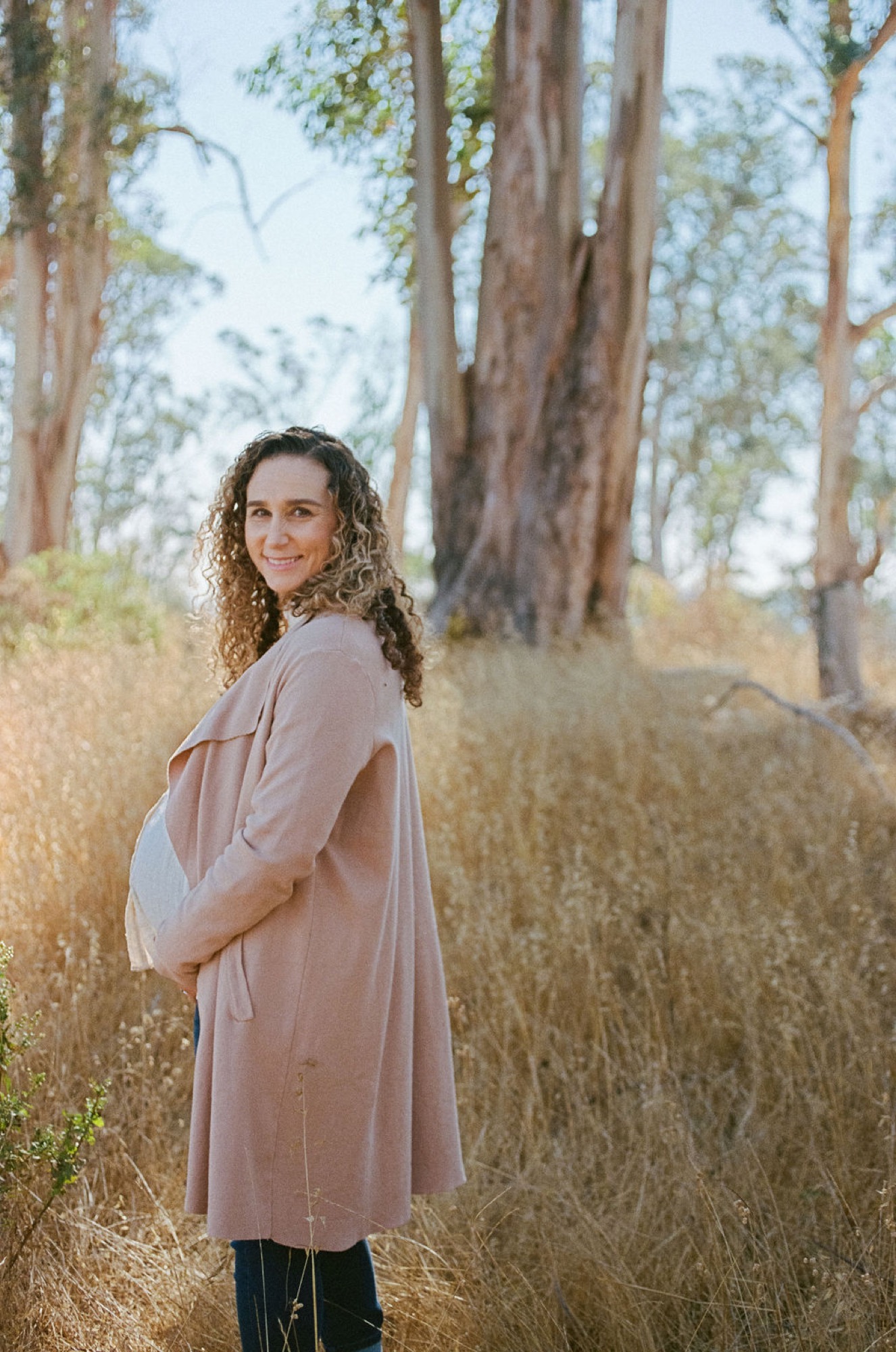 Pregnant woman with curly hair smiles over her shoulder in a sun-dappled eucalyptus grove, wearing a taupe cardigan over white maternity dress