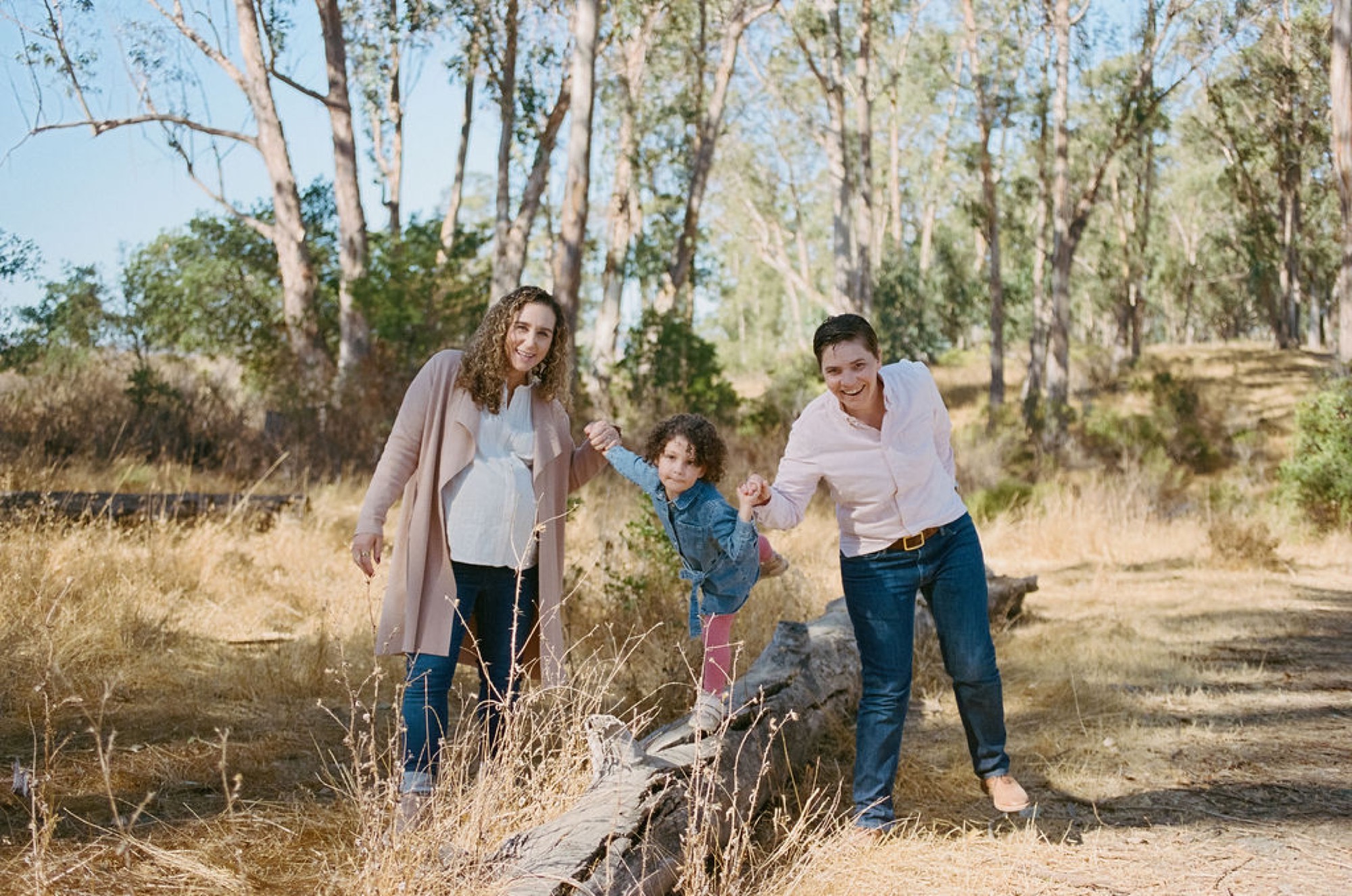 Two women guide a curly-haired toddler along a sun-dappled trail beneath towering eucalyptus trees, their genuine smiles radiating joy