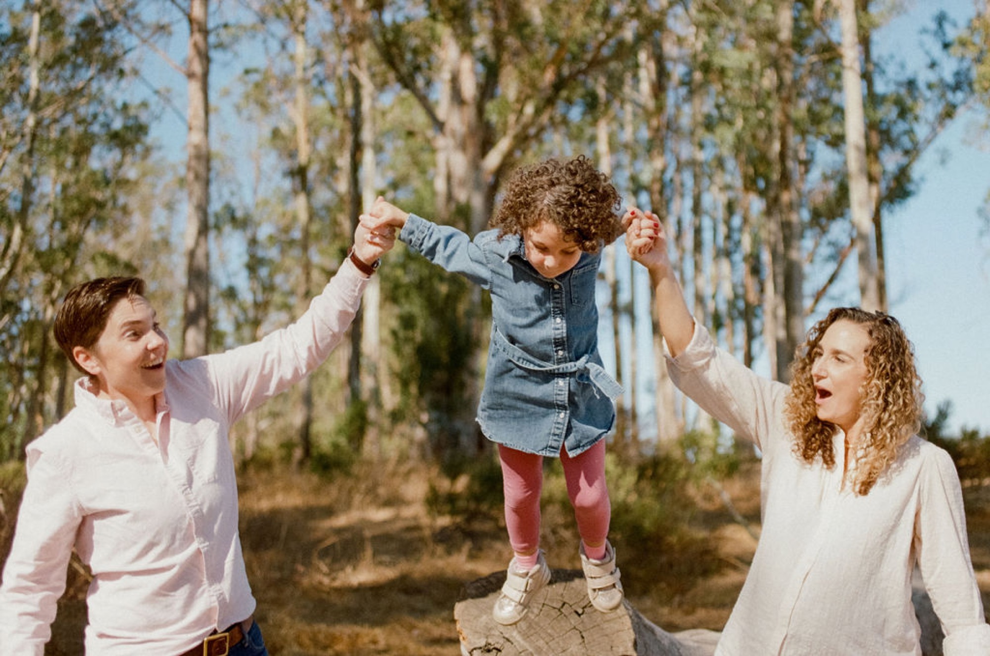 Two women lift their curly-haired toddler between them, all beaming with joy in a sun-dappled forest of tall pines
