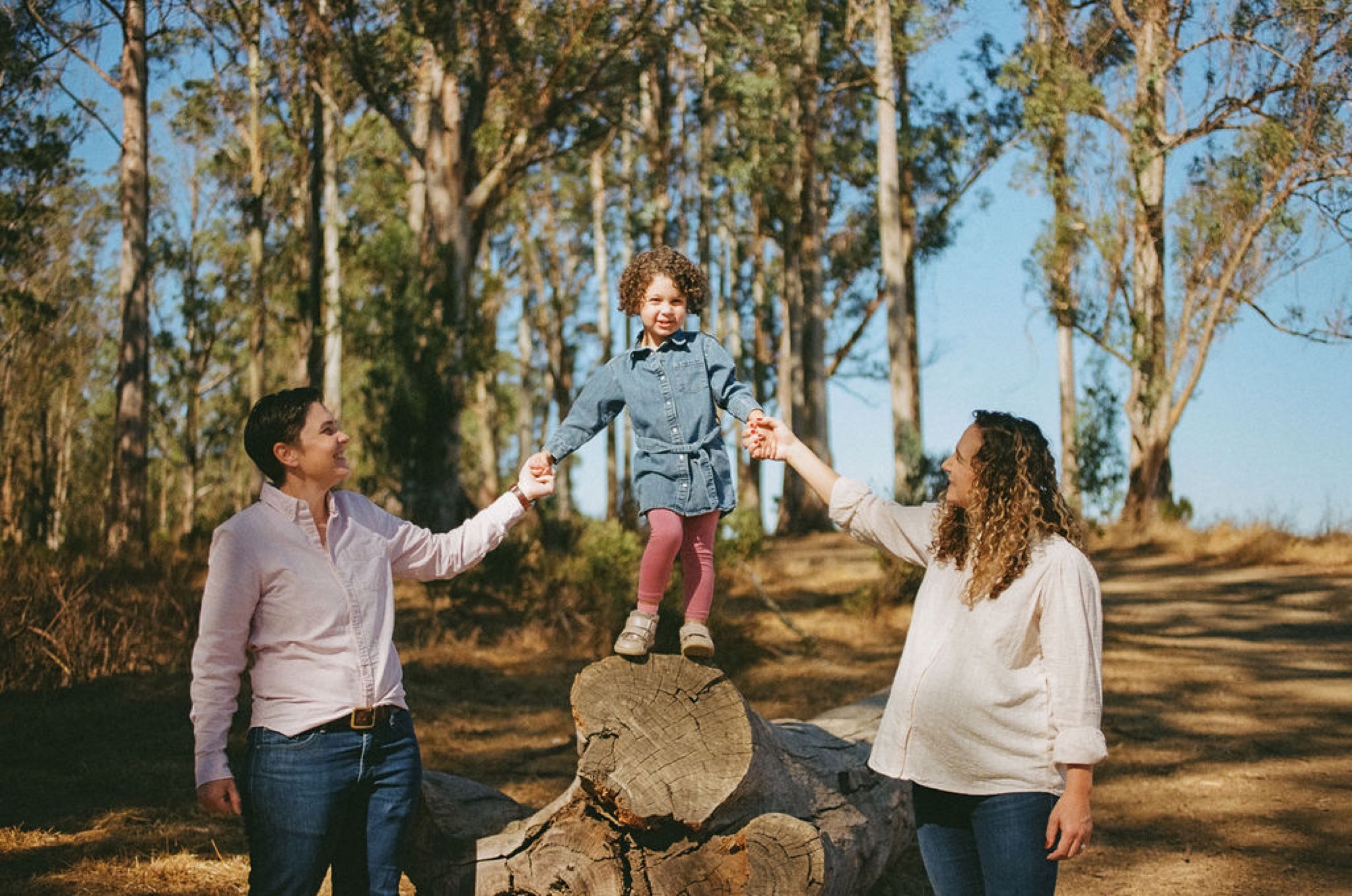 Two parents lift their curly-haired daughter onto a felled tree trunk, her joyful smile radiant against towering eucalyptus trees and dappled afternoon light