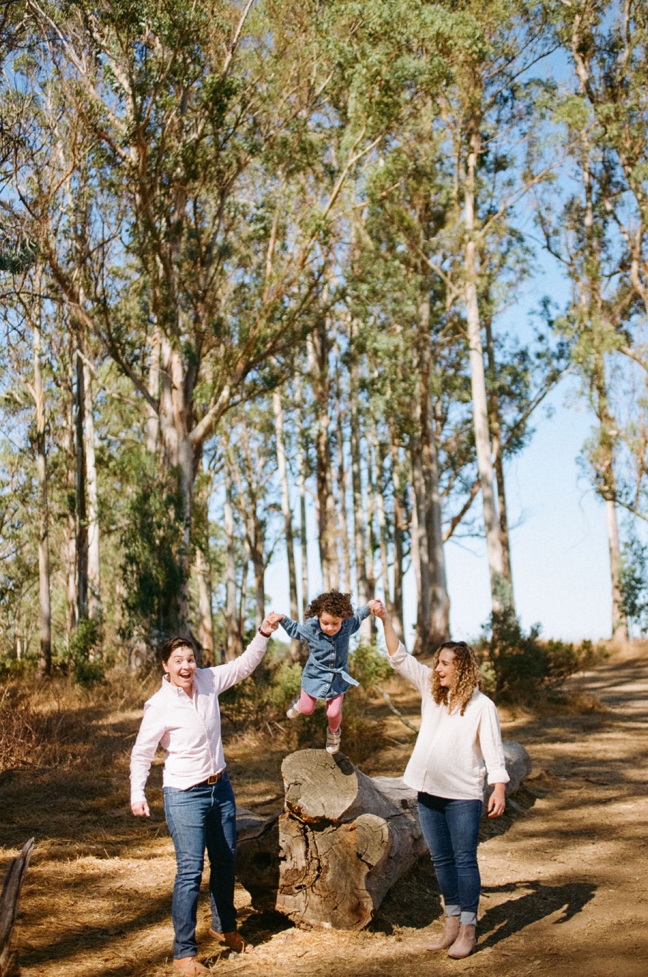 Two parents swing their toddler between them on a sunlit tree-lined path, genuine joy radiating across all three faces