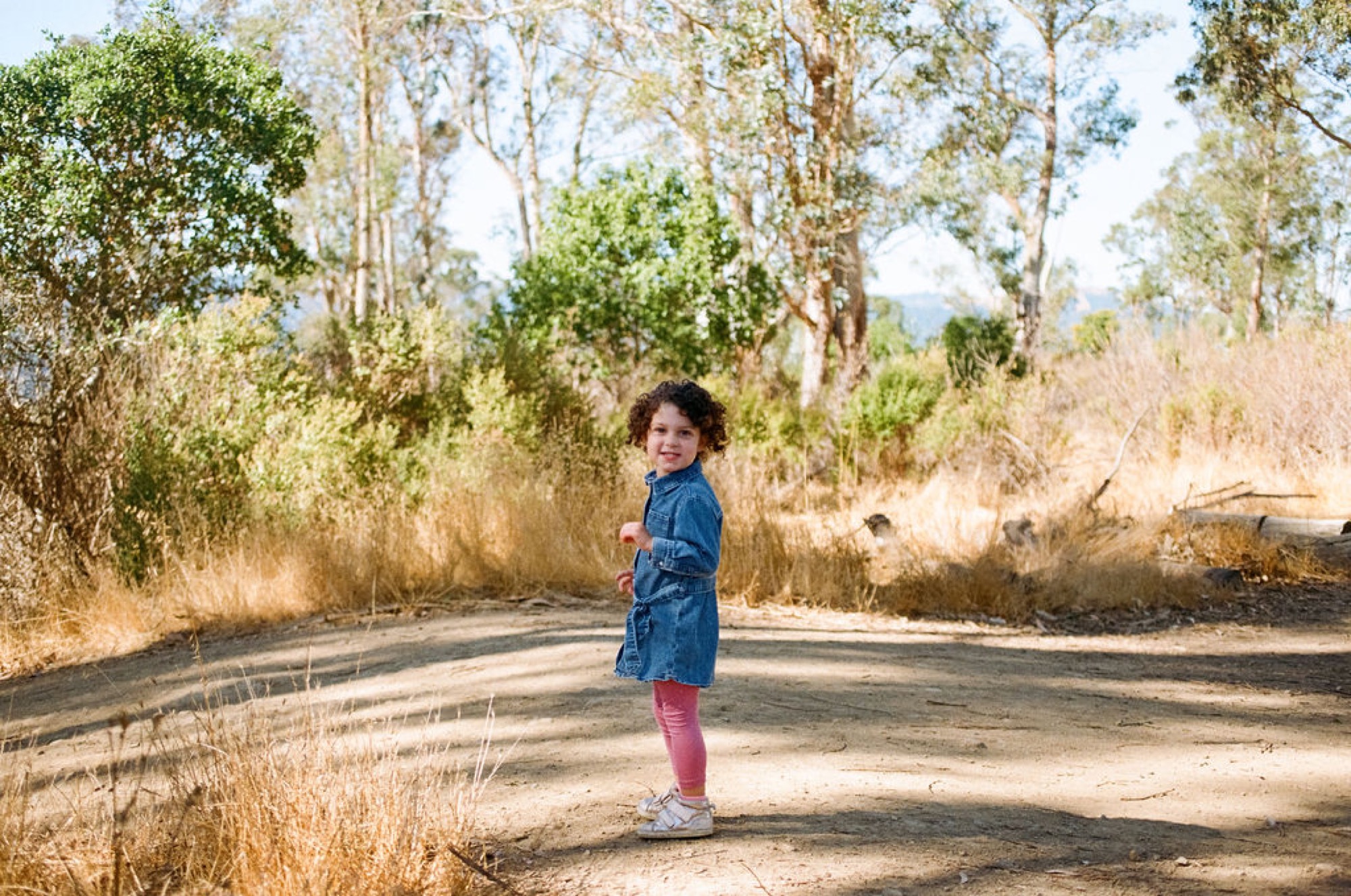 A curly-haired child in denim jogs along a dusty trail, grinning over her shoulder amid tall eucalyptus trees and golden dry grass