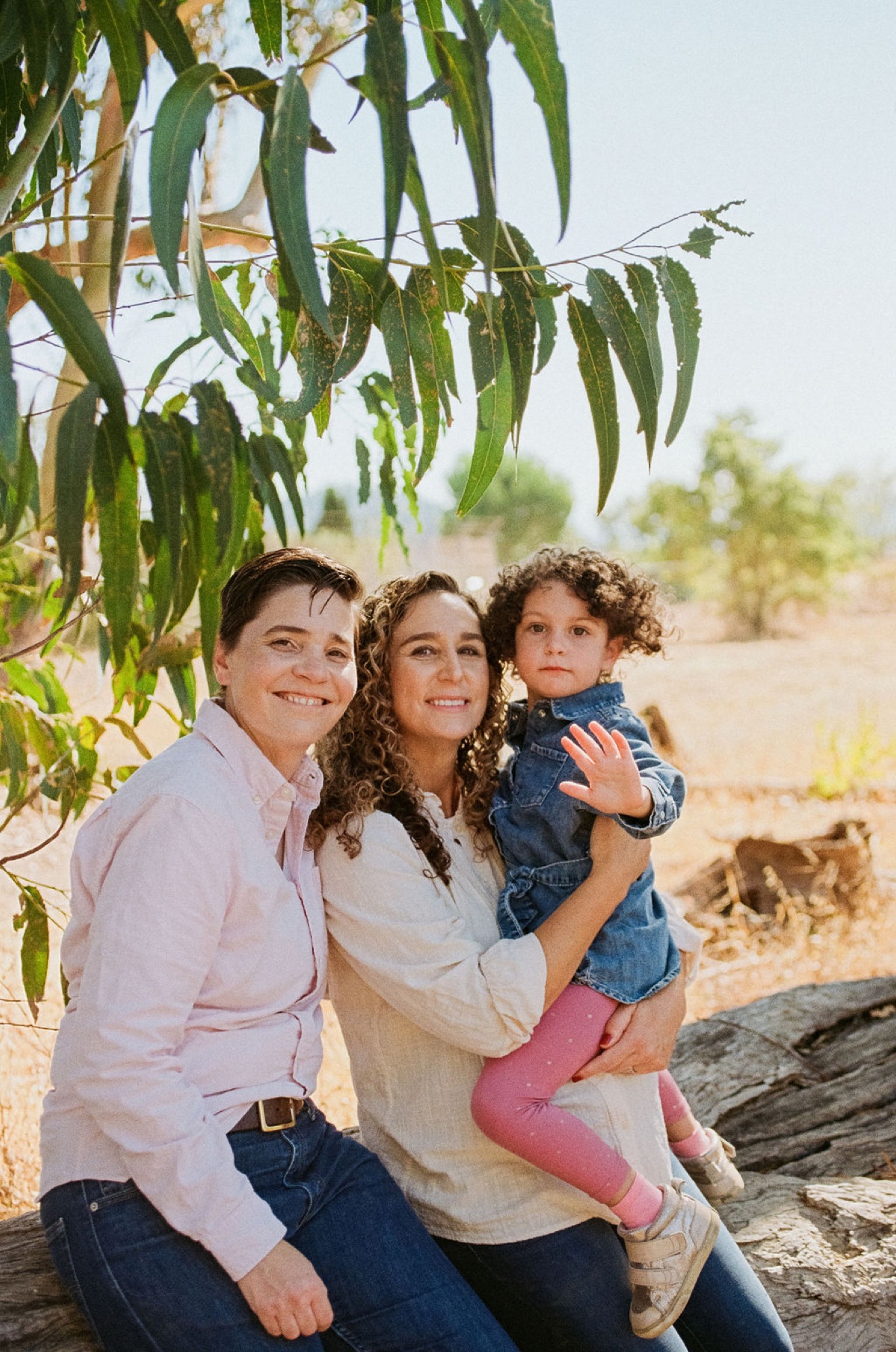 Two adults and a toddler sit together on a log beneath hanging eucalyptus branches, smiling warmly at the camera in golden afternoon light