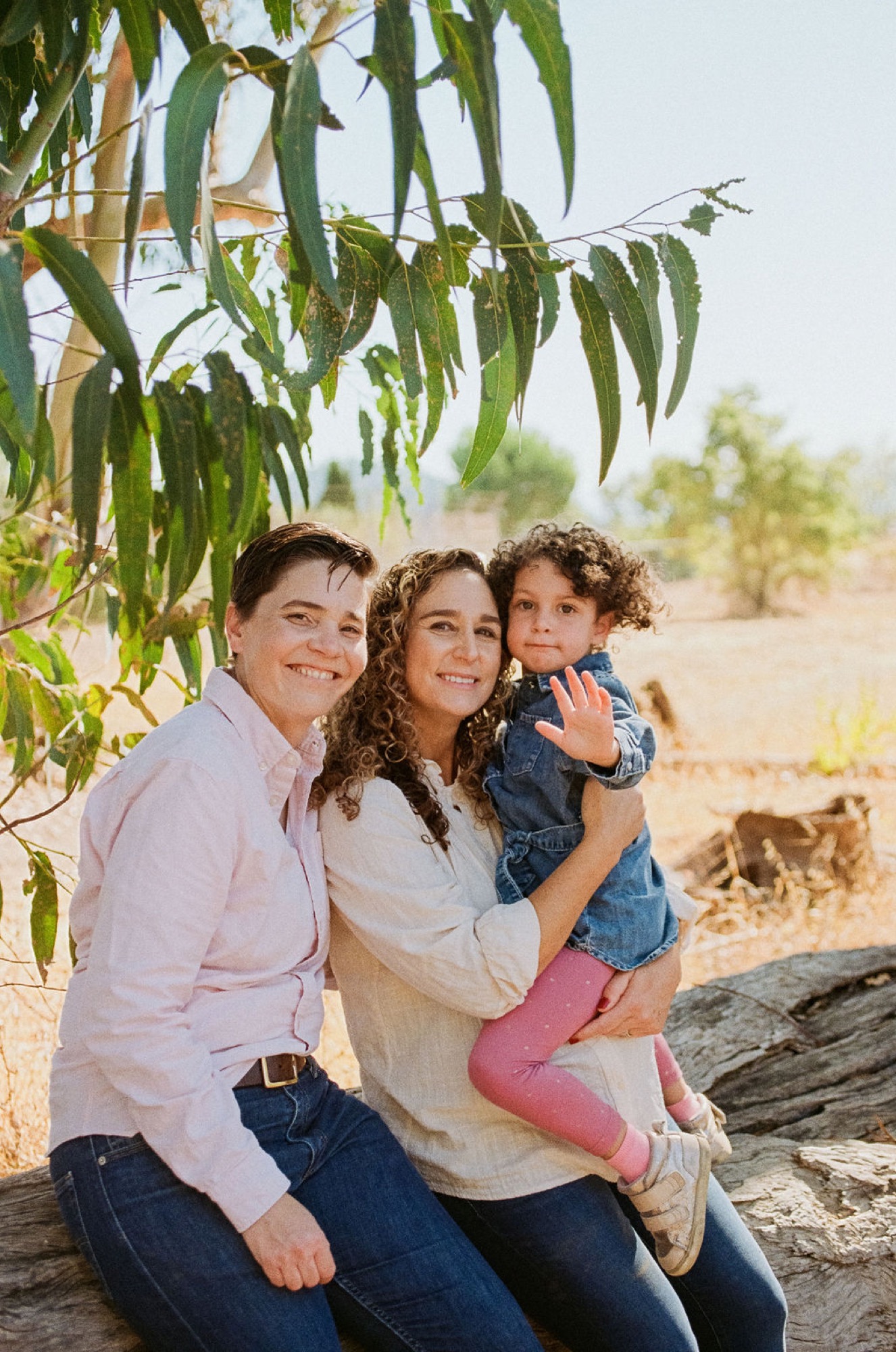 Three siblings sit together on a weathered log beneath drooping eucalyptus branches, their genuine smiles and relaxed poses glowing in warm afternoon light