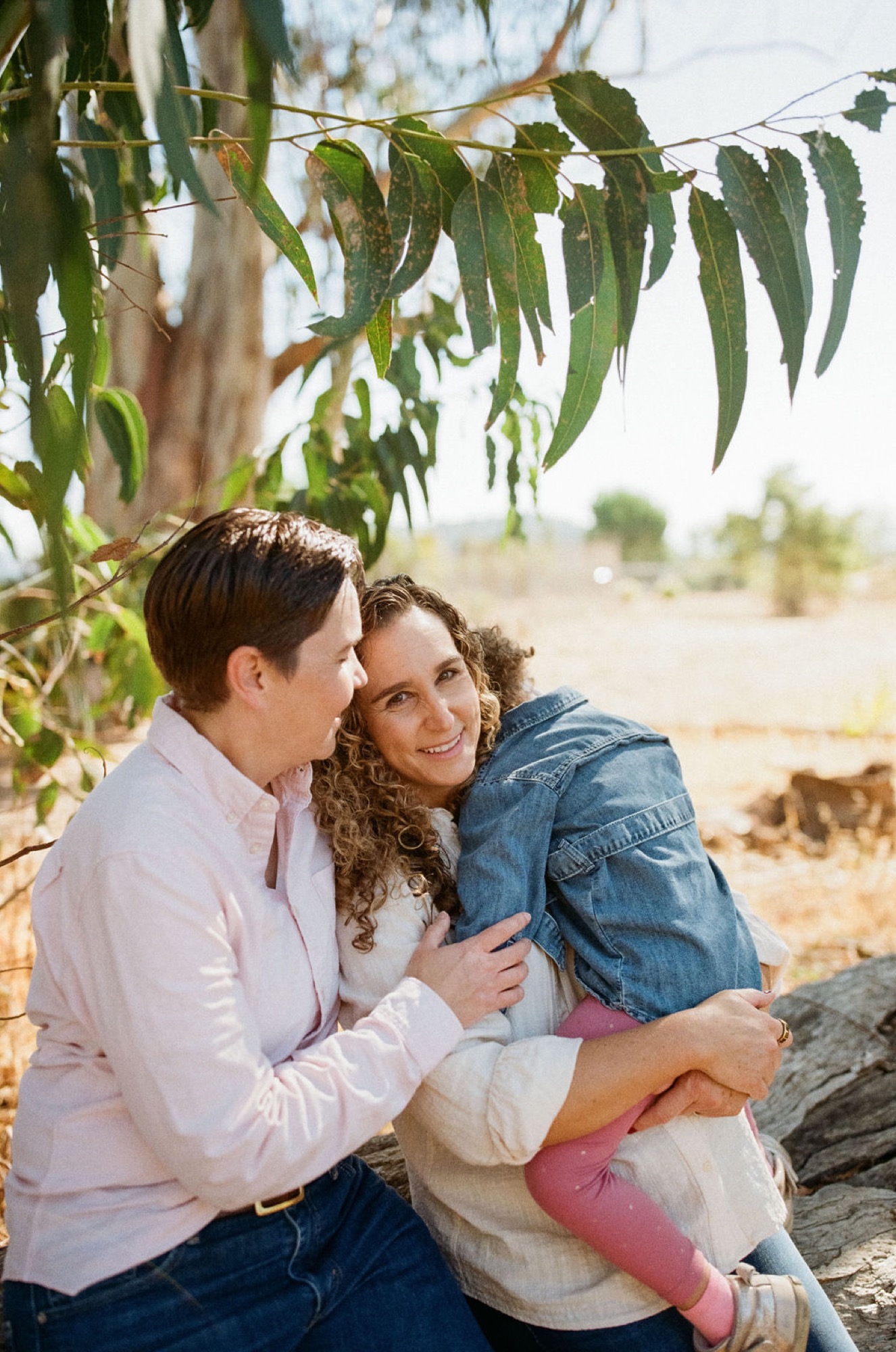 Expectant couple shares an intimate moment beneath hanging willow branches, their genuine smiles and close embrace framed against sun-dappled countryside