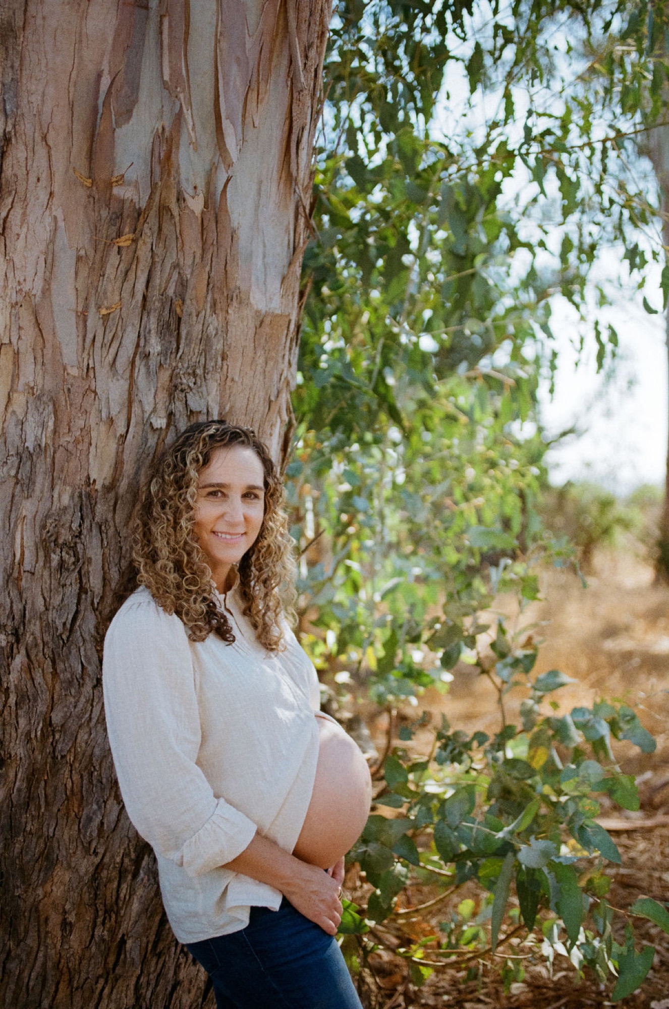 Pregnant woman in white linen leans against weathered eucalyptus trunk, gentle smile, dappled afternoon light filtering through hanging foliage