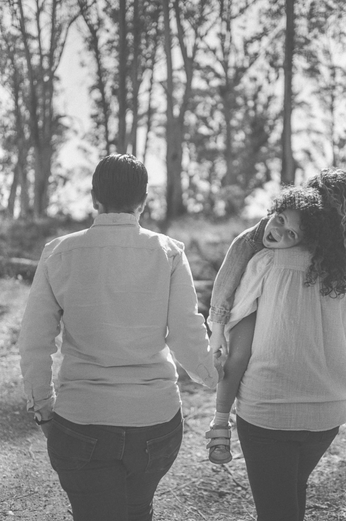 Expectant couple walks hand-in-hand down a sunlit forest path, her joyful expression captured mid-glance toward the camera