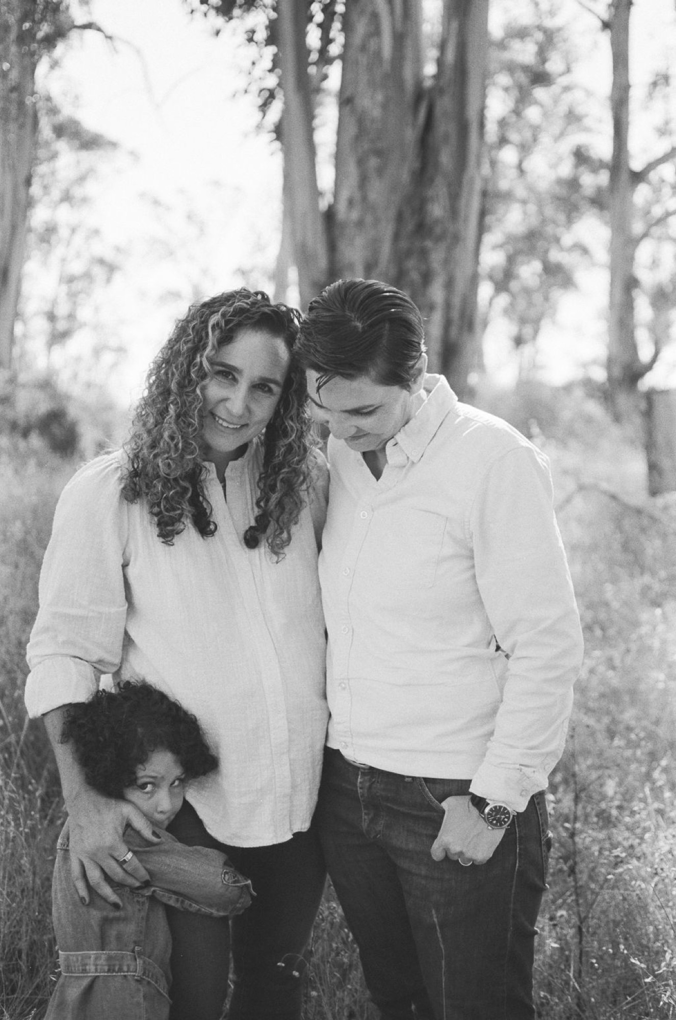 Expectant mother with curly hair smiles warmly while her partner gazes down at their young child between tall redwoods, dappled light filtering through the canopy