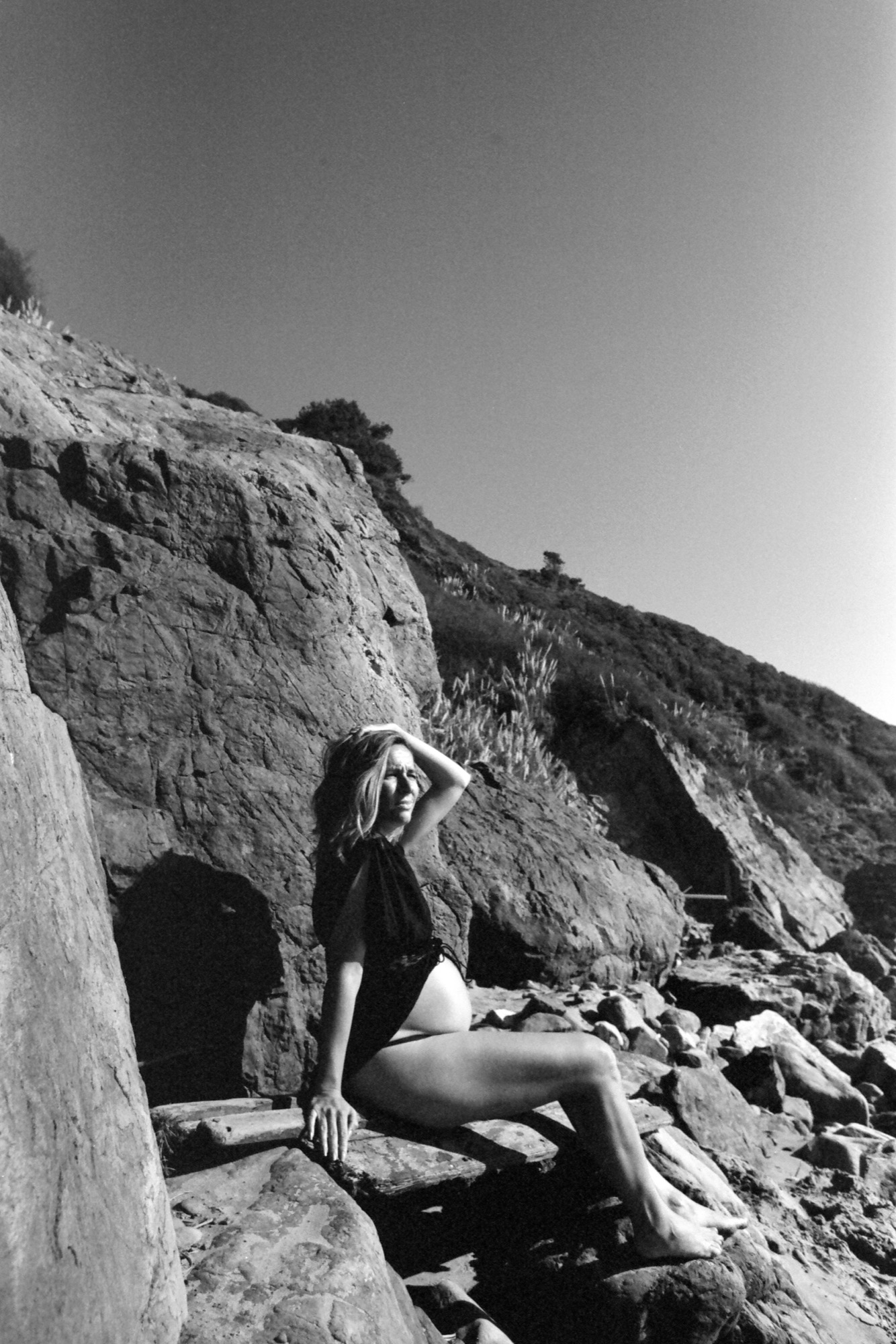 Woman in black shirt and light shorts sits on rocky terrain, hand raised to her head, gazing toward distant hillside vegetation under clear sky