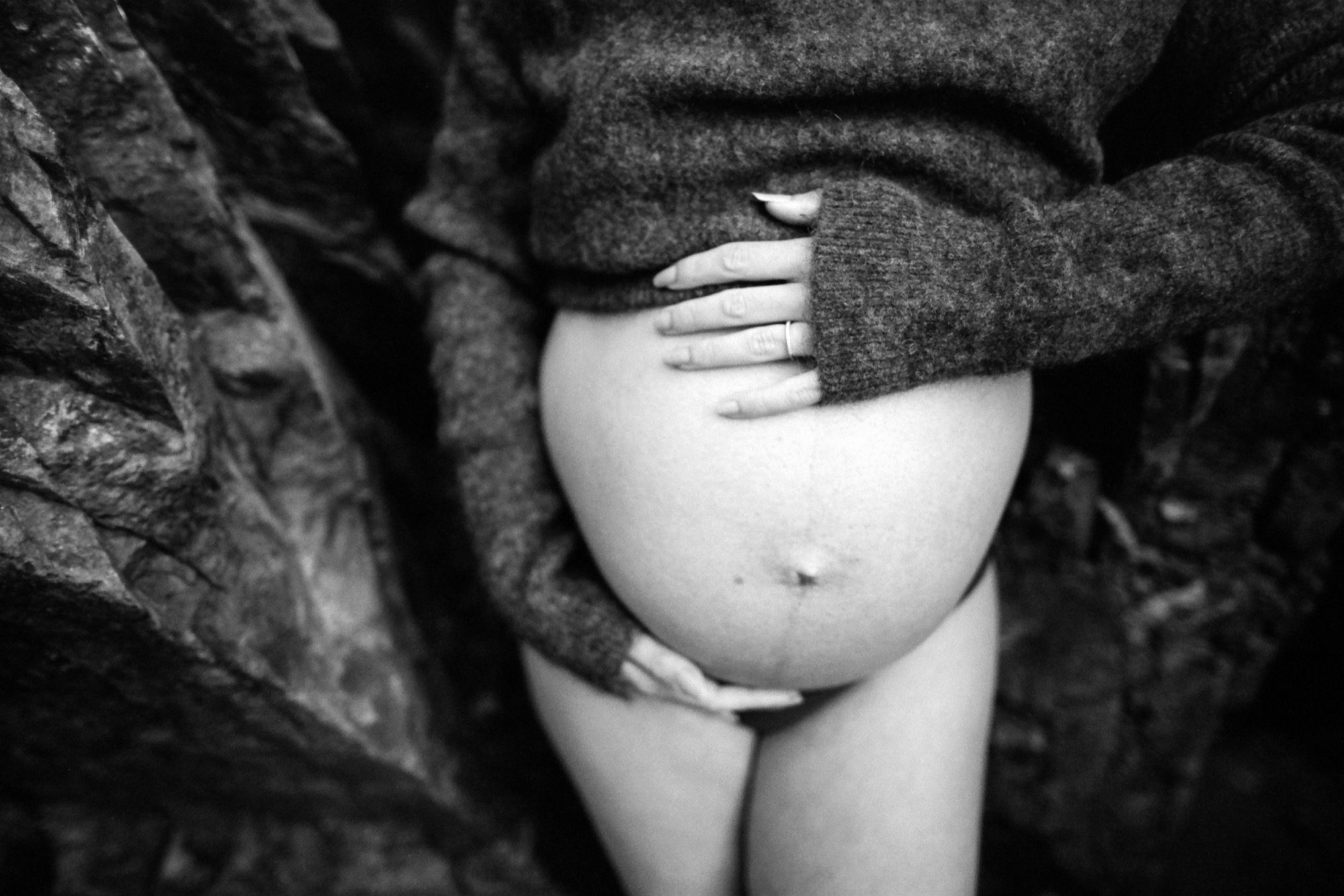 A pregnant woman's bare belly cradled by protective hands, rendered in intimate black and white against layered dark clothing