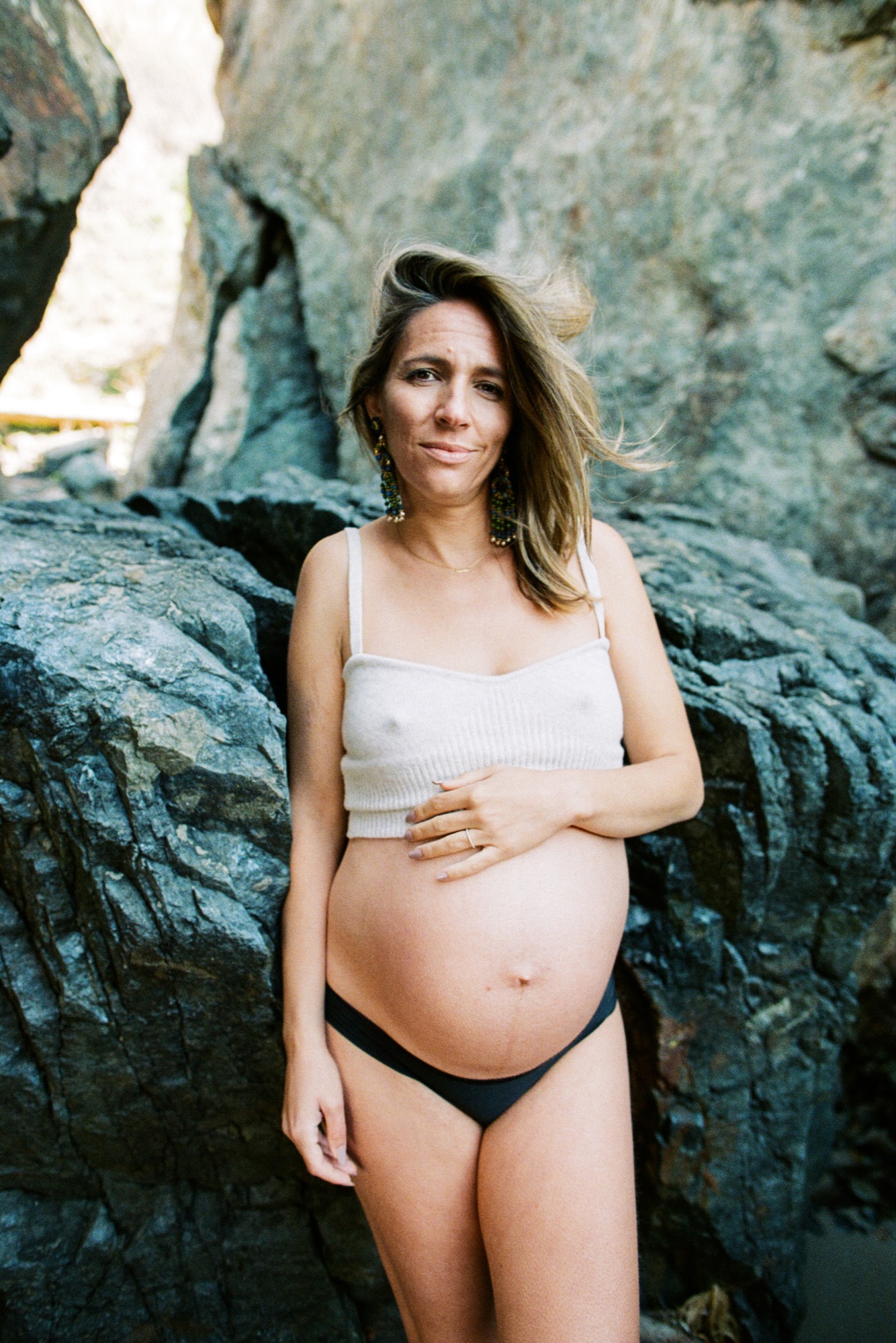Pregnant woman in a white crop top and black bikini bottom poses between towering granite boulders, one hand cradling her belly, golden light filtering through the rocks