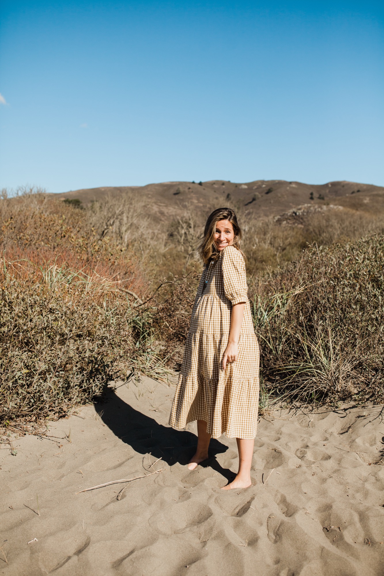 Pregnant woman in cream gingham dress stands barefoot on sand, turning toward camera with warm smile as golden afternoon light casts her shadow across the beach