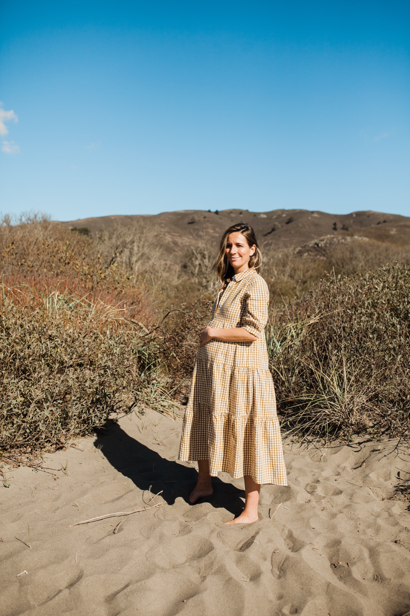 Pregnant woman in cream gingham dress stands barefoot on sand, gazing over her shoulder with gentle expression as rolling hills and dry brush rise behind her under clear blue sky