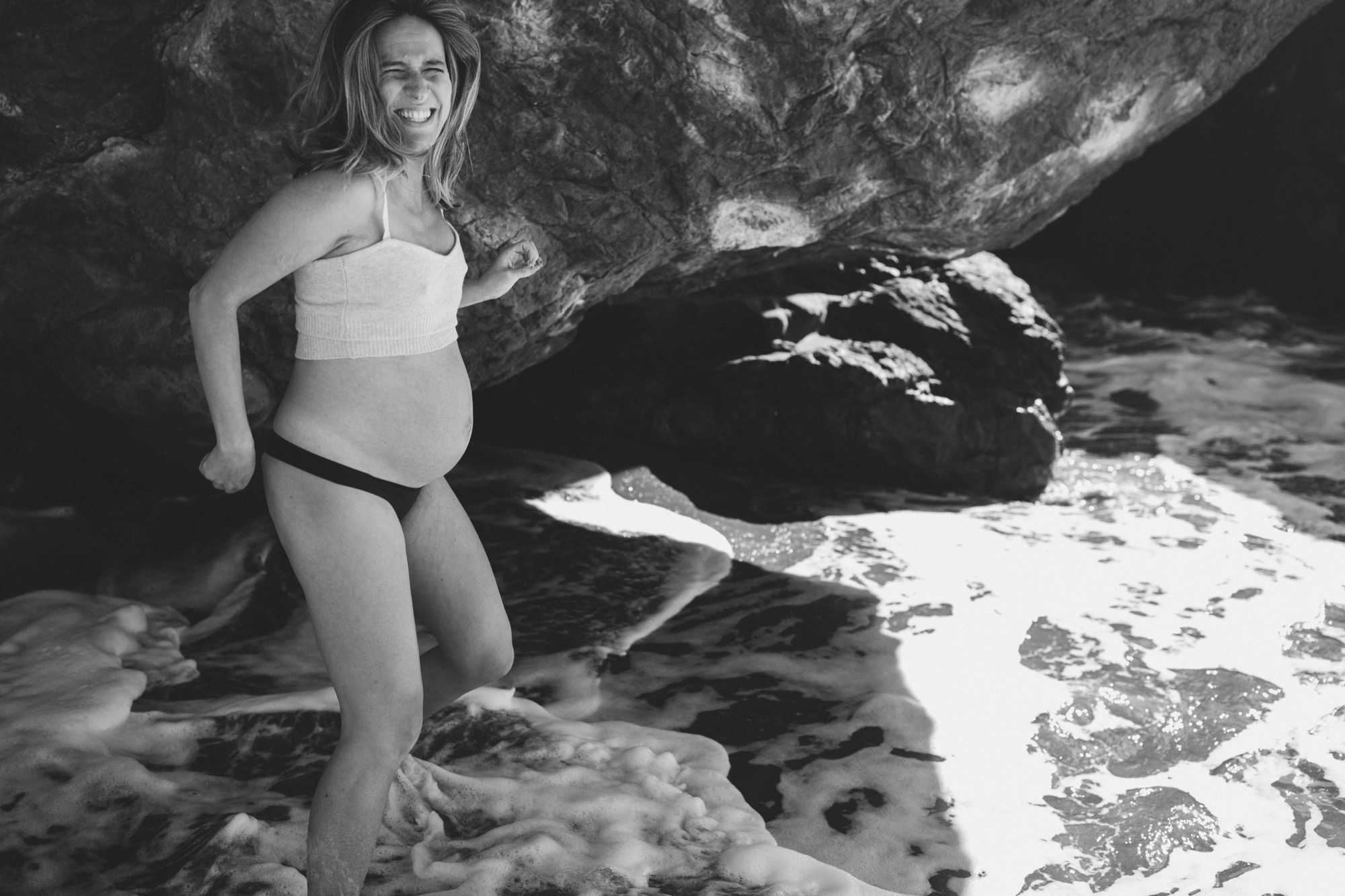 Pregnant woman smiles radiantly at rocky beach cove, white crop top and dark bikini bottom, dramatic rock formations and foaming surf behind her