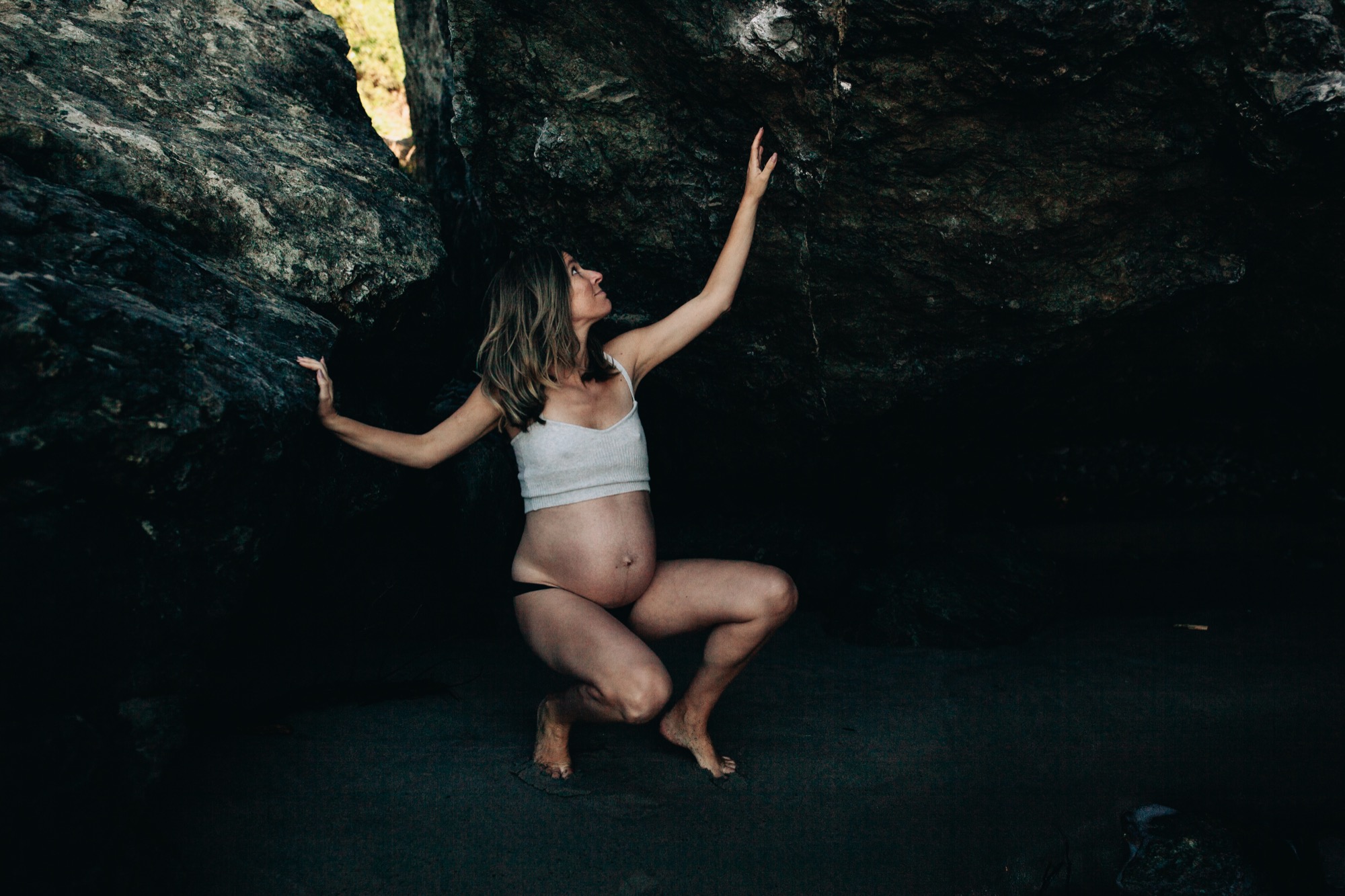 Pregnant woman crouches barefoot against dark cave rocks, one arm reaching upward toward golden light filtering through the opening above