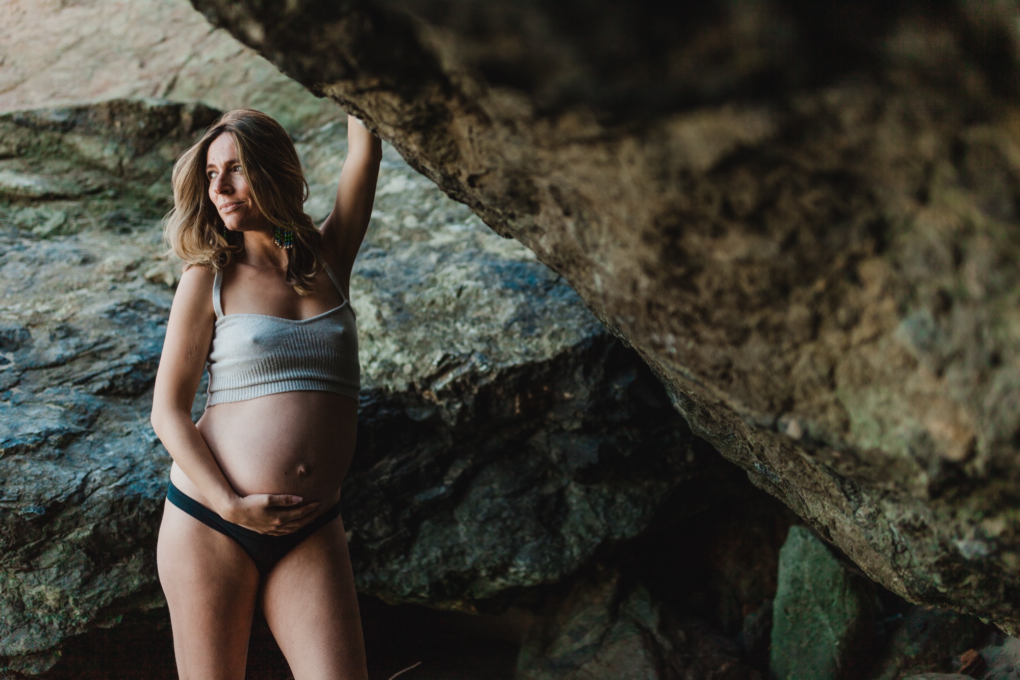 Pregnant woman in light blue crop top gazes upward while touching moss-covered rock overhang, golden sunlight illuminating her face and belly