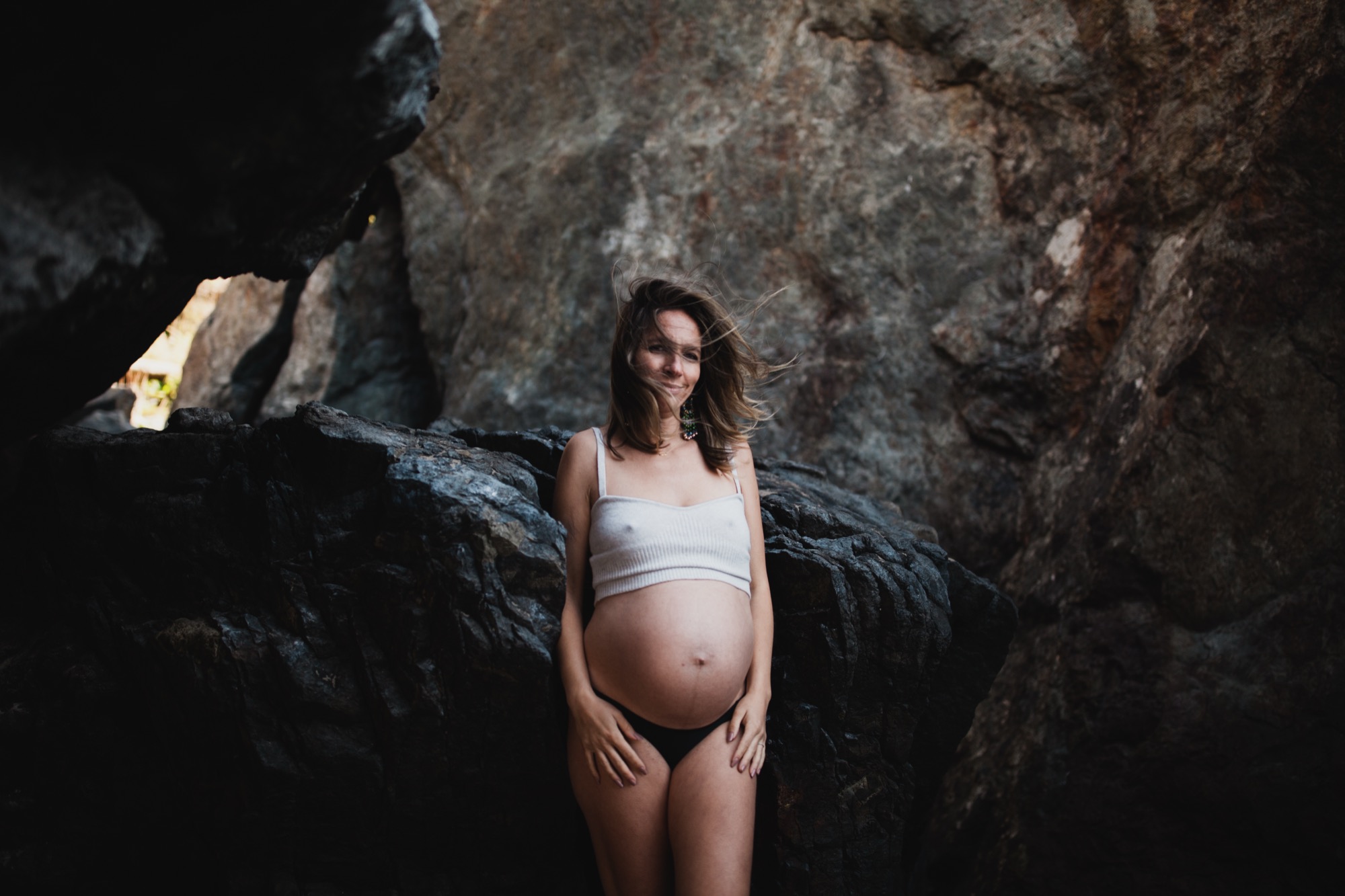 Pregnant woman in white crop top stands smiling between dark rocky outcroppings with warm golden light filtering through the stone crevice behind her