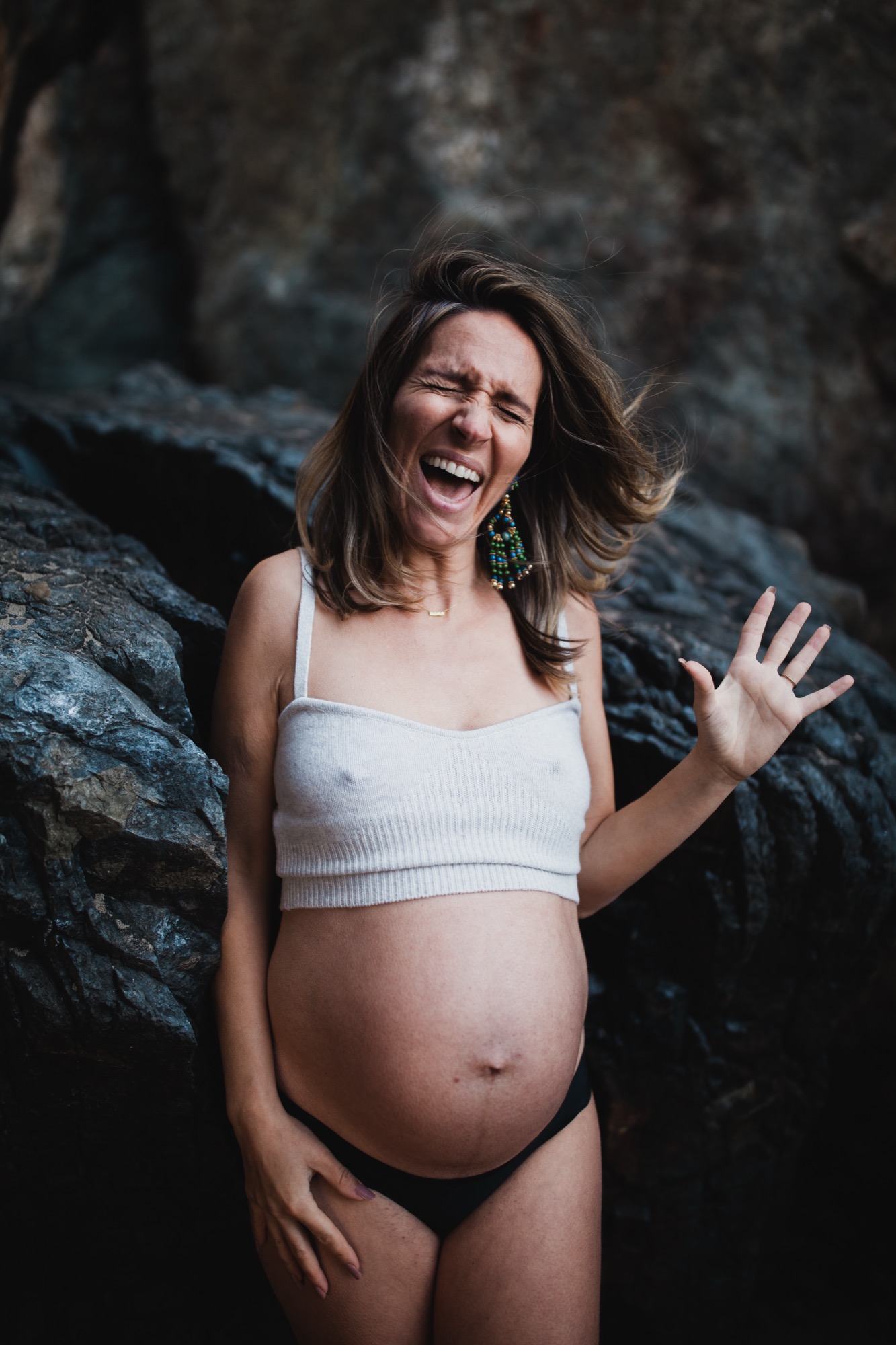 Pregnant woman laughs joyfully against dark coastal rocks, one hand raised expressively, wearing white crop top and beaded earrings