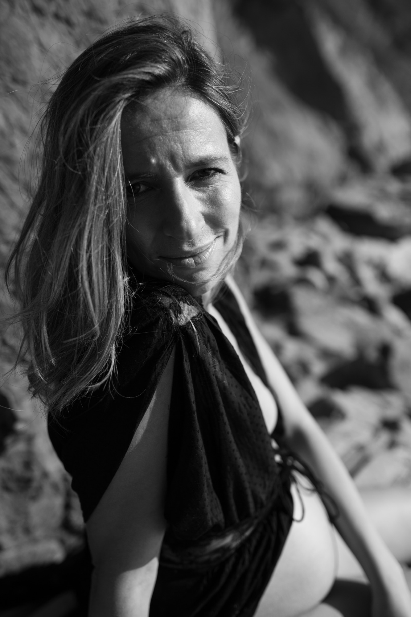 Pregnant woman gazes downward with serene expression, long dark hair flowing across her shoulder in soft natural light against rocky terrain