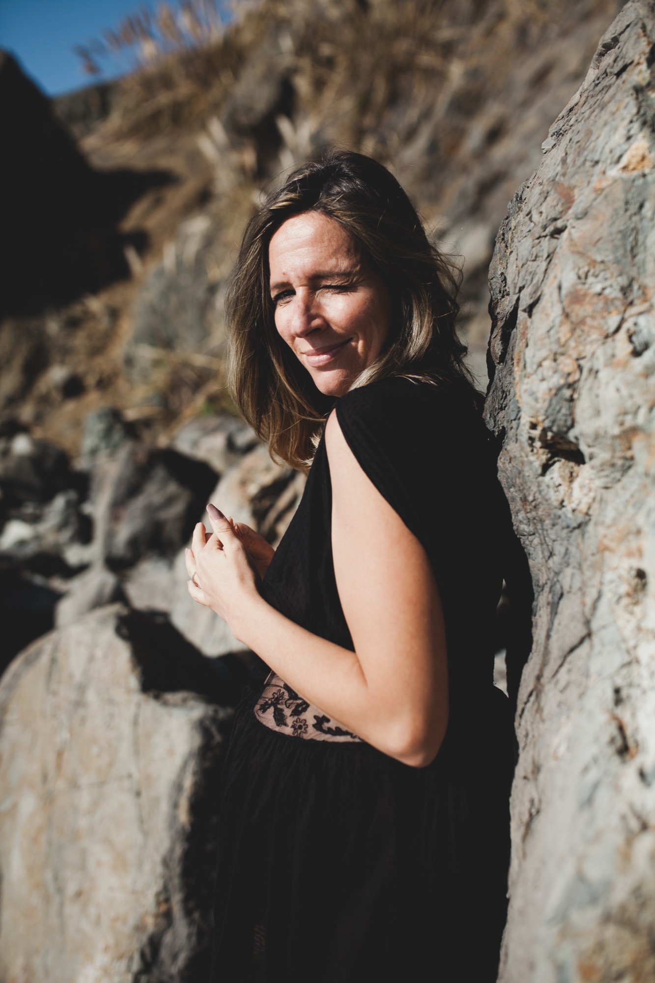 Pregnant woman in black sleeveless dress leans against weathered stone wall, gazing over shoulder with gentle smile in bright coastal sunlight