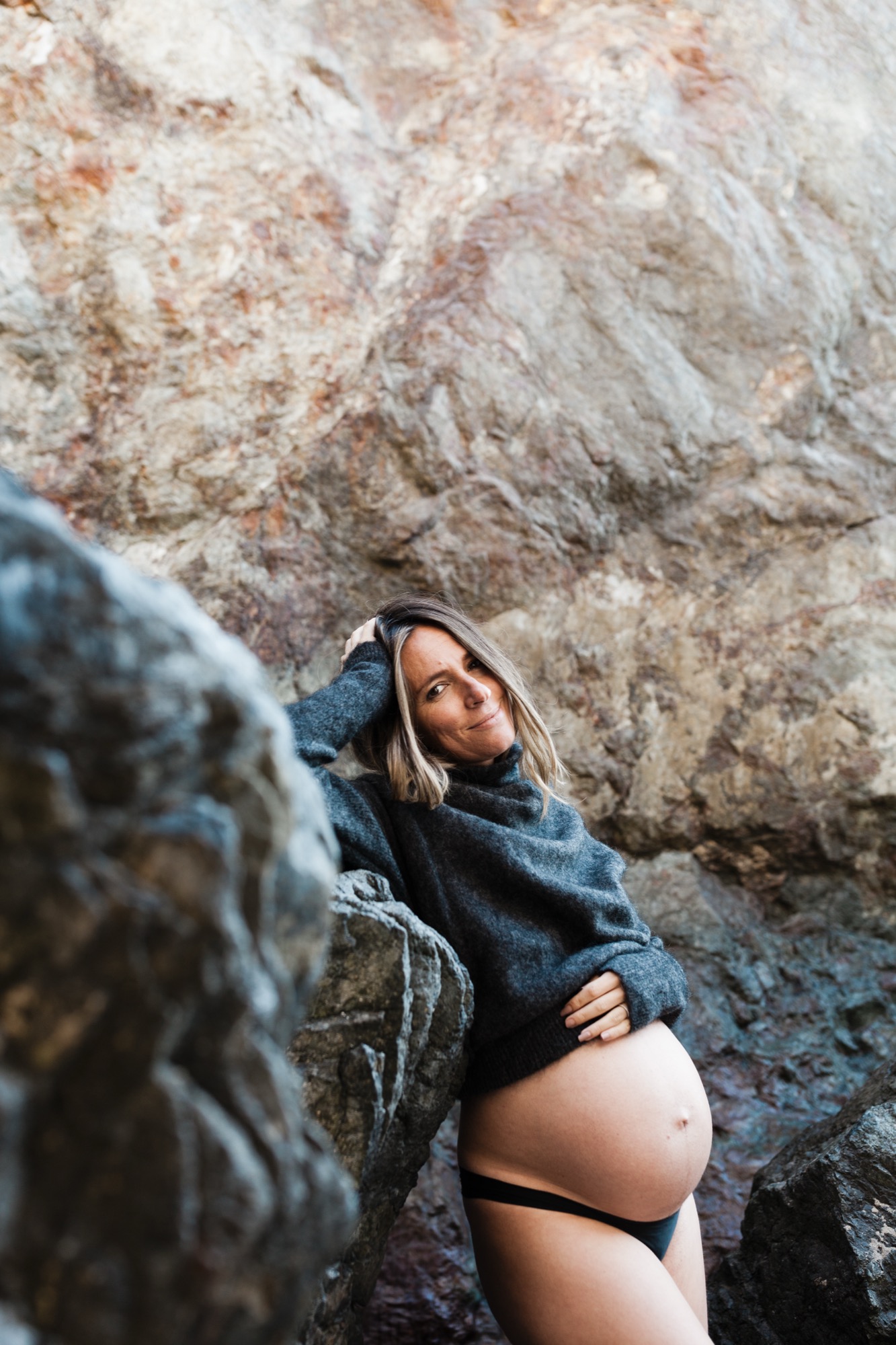 Pregnant woman in charcoal sweater gazes upward between weathered rock formations, hand cradling her belly with serene confidence