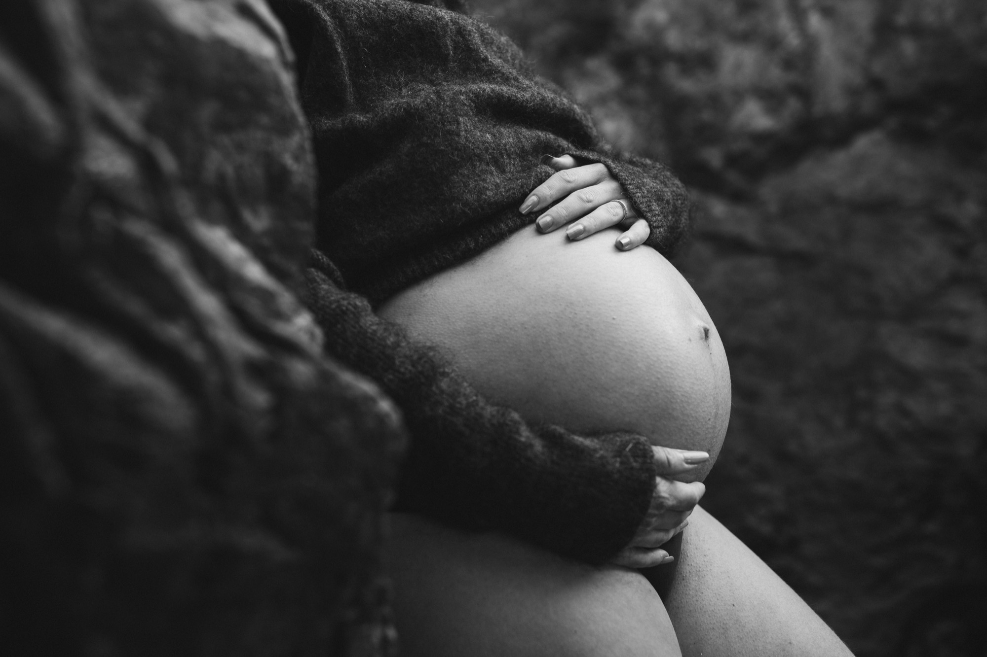 A pregnant woman in a dark sweater cradles her belly while small hands rest gently on her rounded abdomen in intimate black and white portraiture