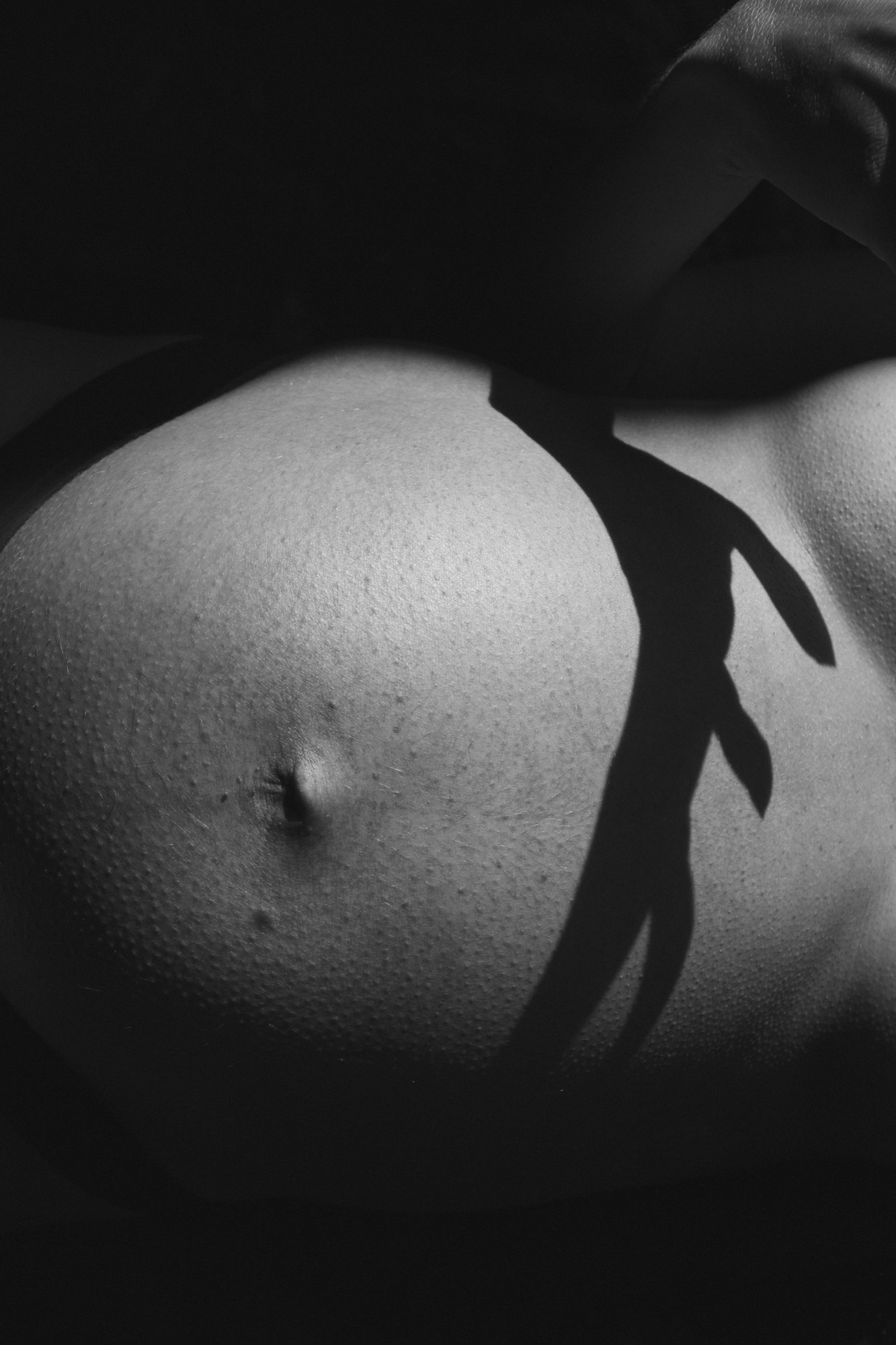 Dramatic black and white maternity portrait with side lighting emphasizing the curve of pregnancy against deep shadows