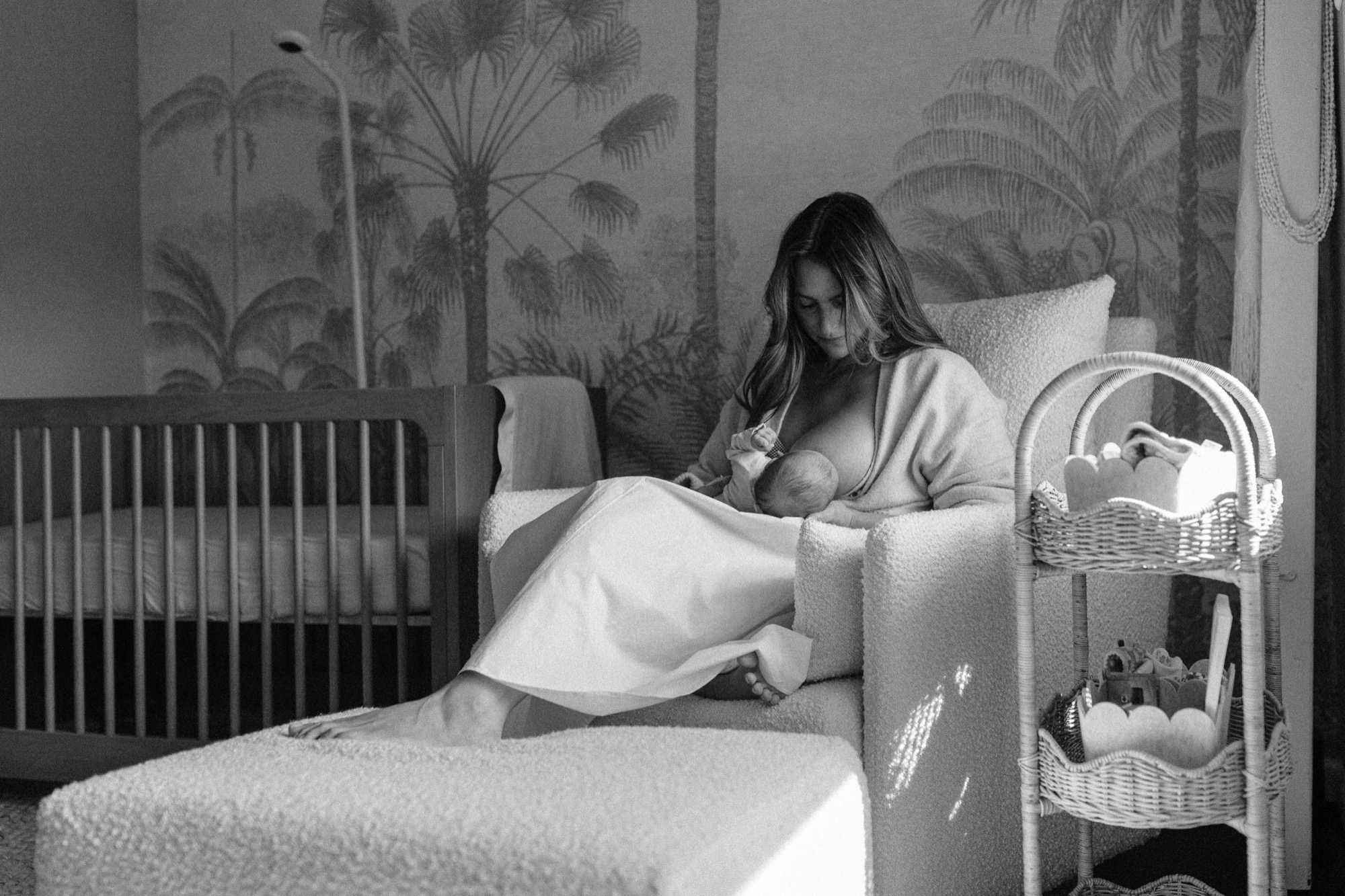 A quiet moment of stillness in her Marin nursery — soft window light washing over her as she nurses her newborn in the boucle chair, the tropical wallpaper and warm shadows making it feel like the rest of the world simply paused. This is exactly the kind of real, unhurried moment that makes lifestyle newborn photography so worth it — San Anselmo Newborn Photographer