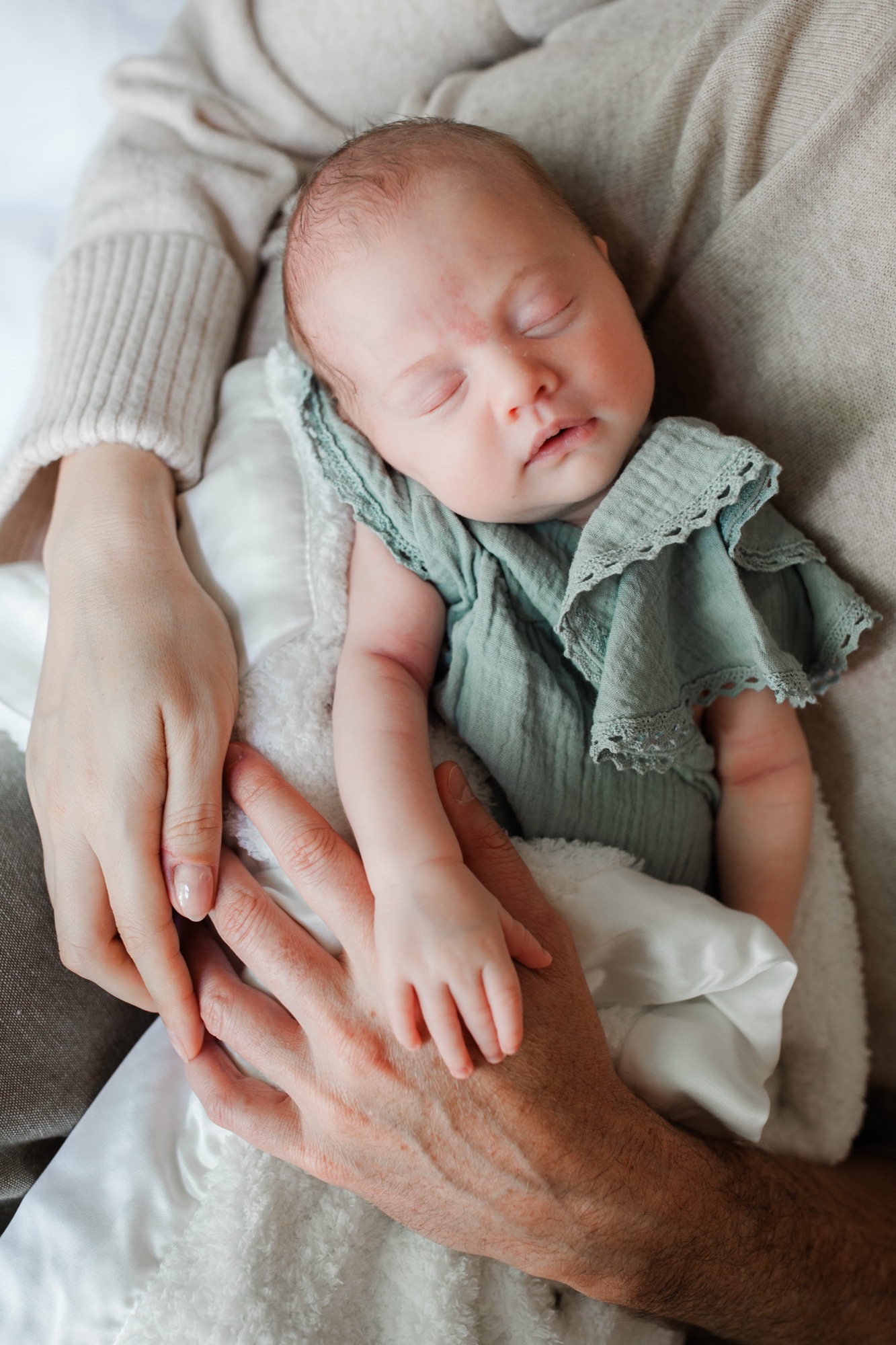 A sleeping newborn in a soft mint lace romper, cradled gently between two pairs of loving hands — her tiny arm reaching outward as if already finding her people. Captured in the quiet of their Marin County home, this moment of shared parenthood is exactly the kind of connection that makes newborn photography so meaningful.