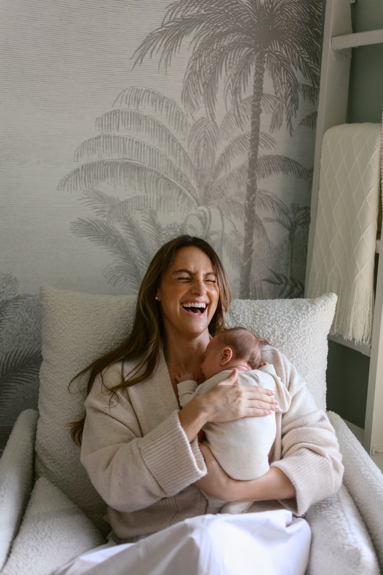 A mother laughs while cradling her newborn close in soft window light