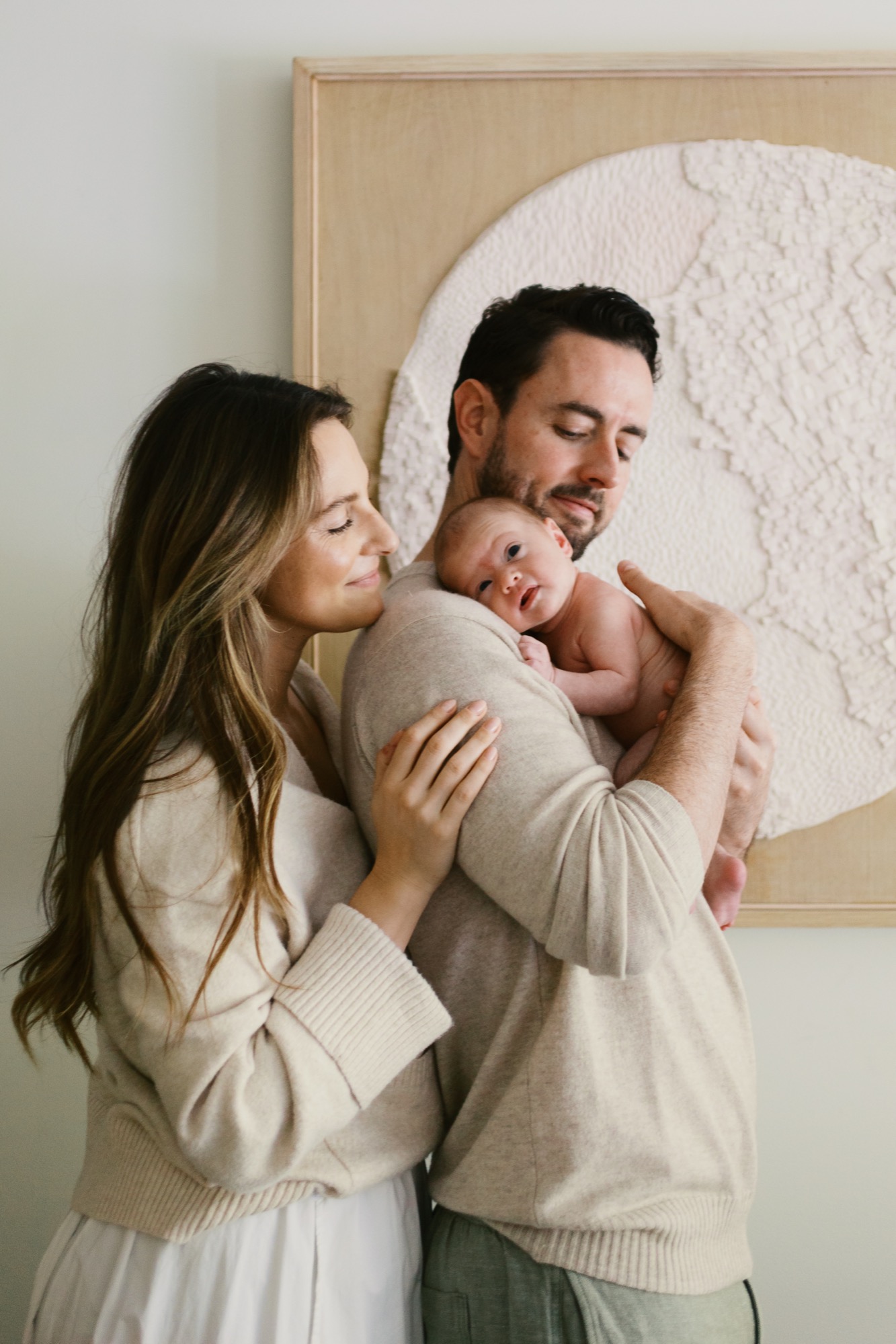 Bathed in soft window light at their Marin County home, this new mom and dad lean in close around their alert, wide-eyed newborn — mom with a tender kiss to baby's cheek, dad cradling them with a quiet, protective smile. It's the kind of unguarded moment that makes you feel the whole world just got a little smaller and sweeter.