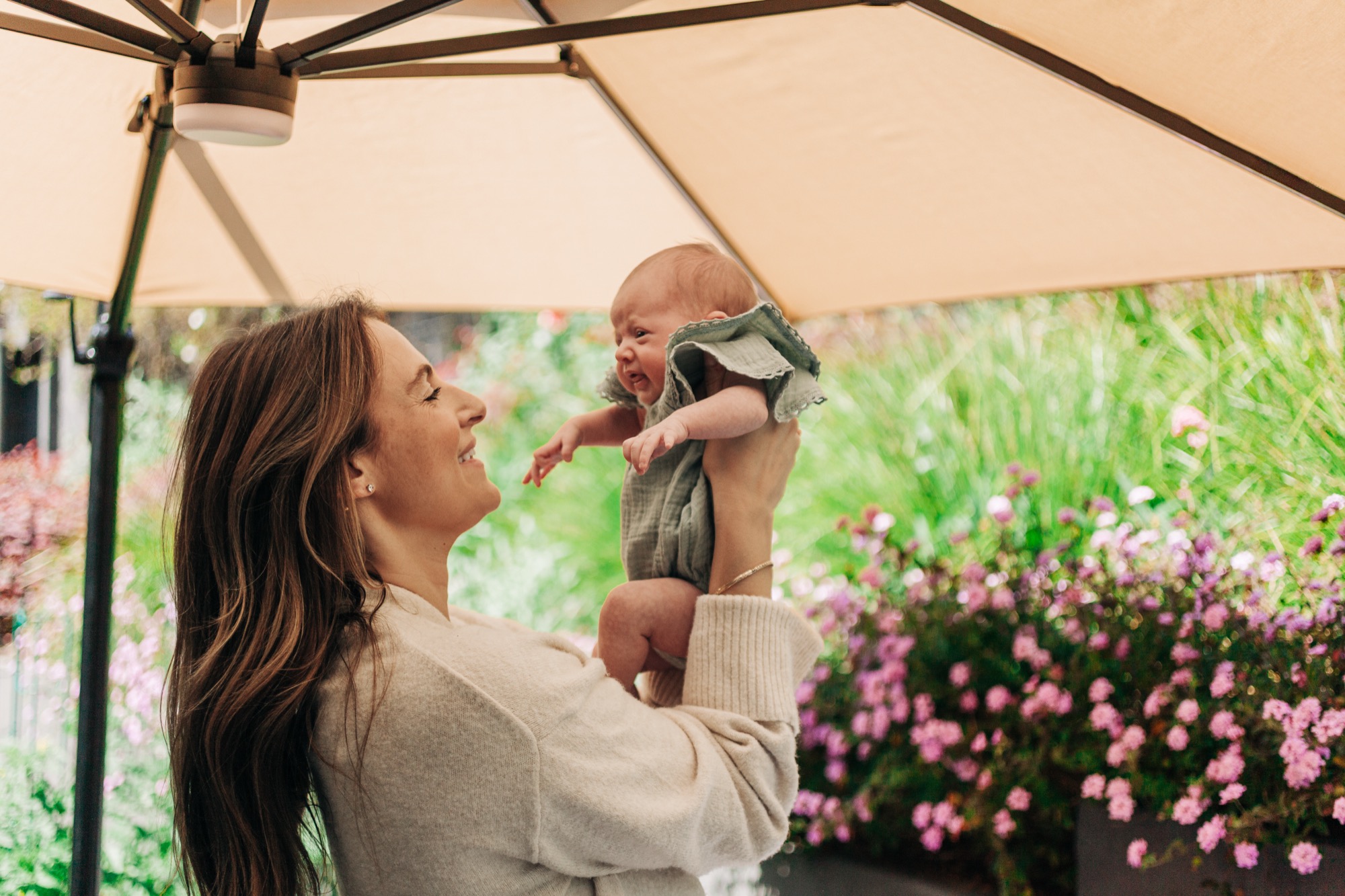 A mother lifts her newborn overhead in a sun-dappled garden