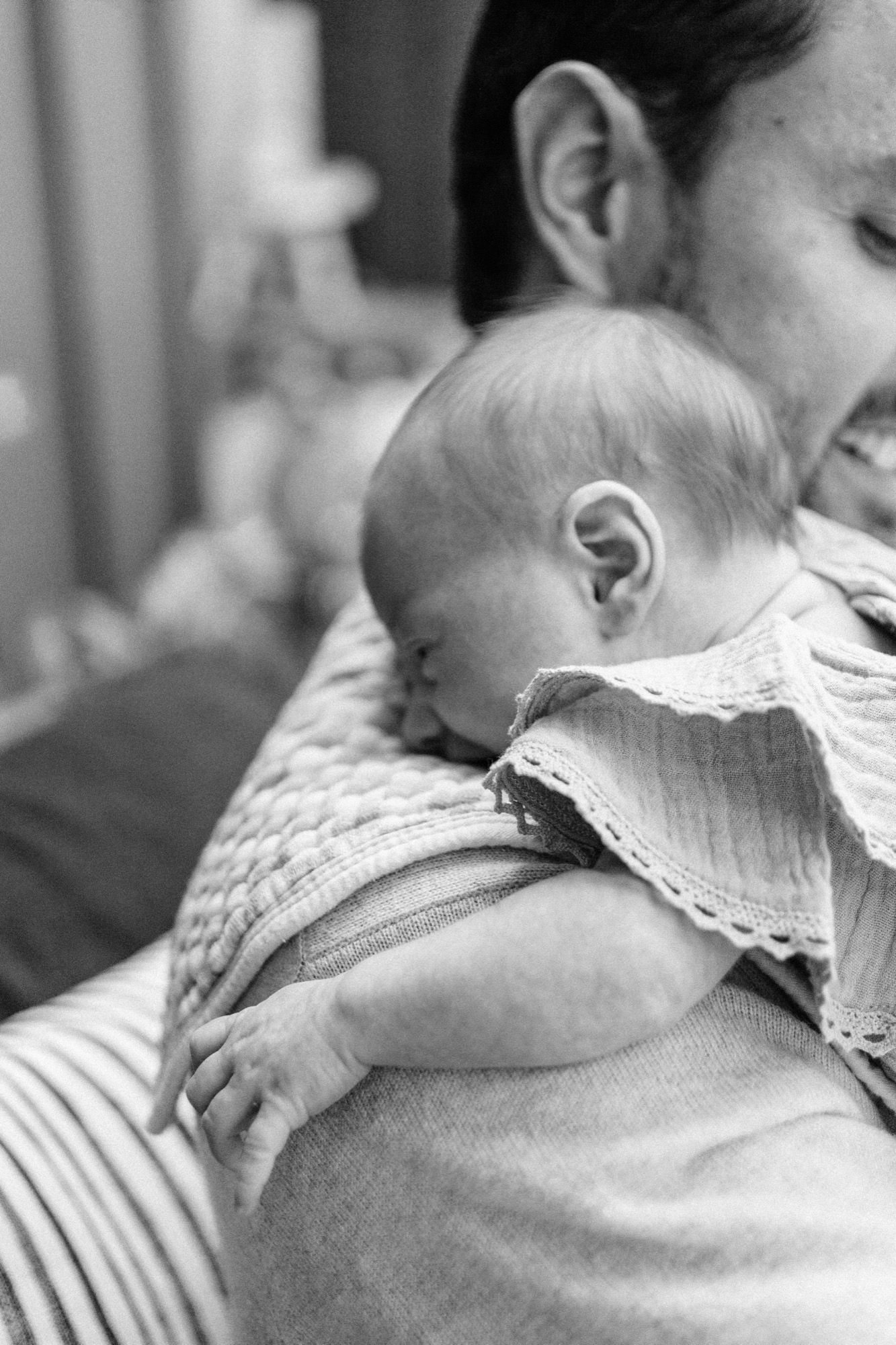 A sleeping newborn rests completely at peace on dad's shoulder, one tiny hand hanging loose and a soft chunky knit tucked close — captured in the warm natural light of their Marin County home. Dad's gentle smile glows quietly in the background, saying everything about what this moment means.