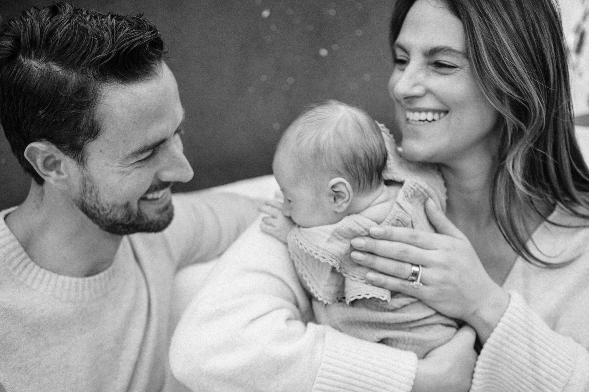 A brand-new family in their Marin County home, wrapped in cream and sage, completely lost in their own little world — mom laughing with pure joy, dad gazing quietly down at their newborn, and one tiny fist curled upward as if reaching toward all that love.