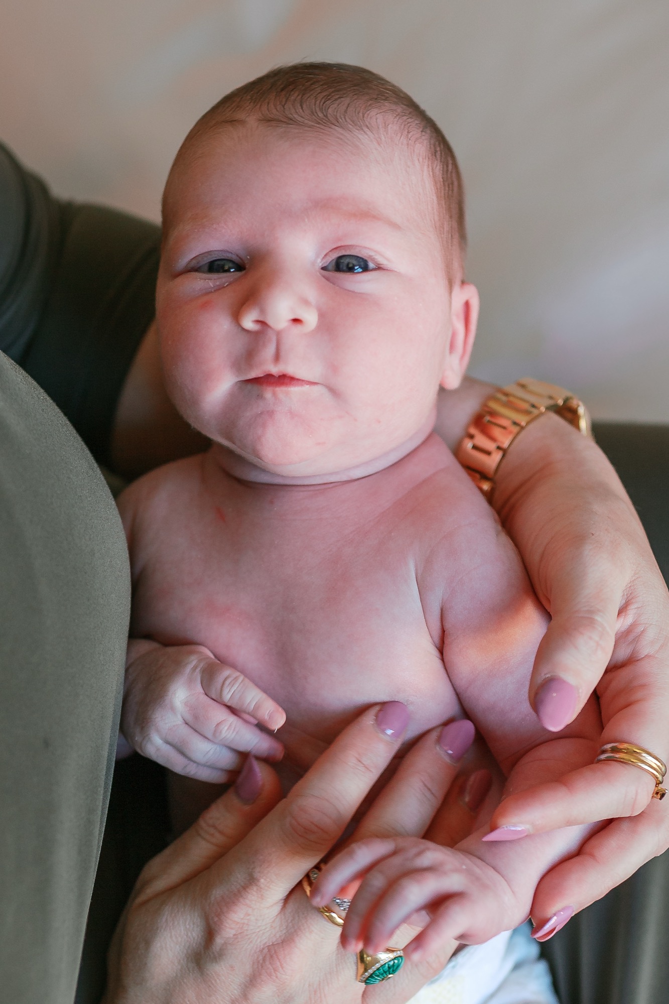 Newborn cradled in parent's hands with alert gaze, soft natural light illuminating delicate features and gold jewelry