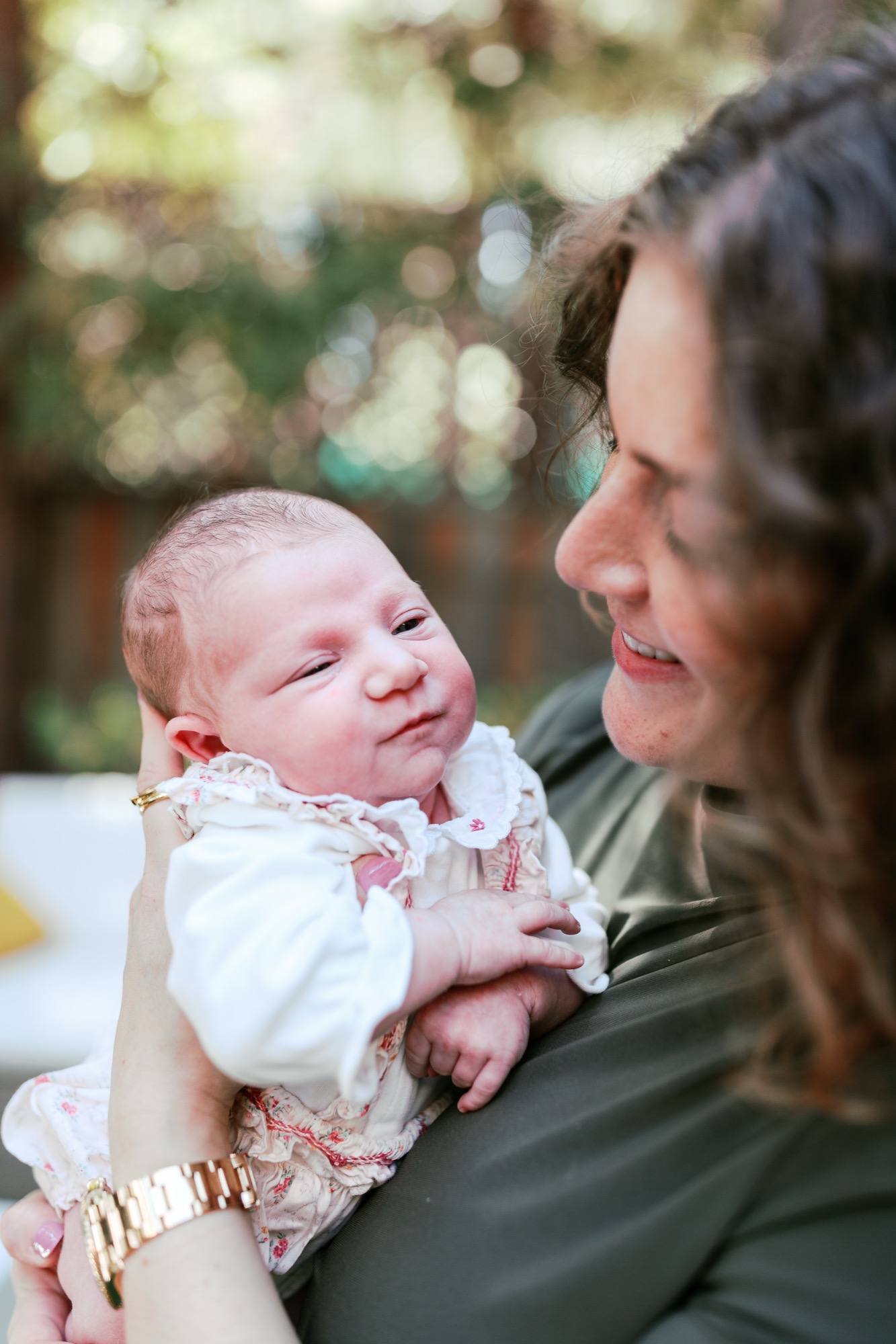 A mother holds her newborn daughter with a genuine smile as soft afternoon light catches their profile against a blurred garden backdrop
